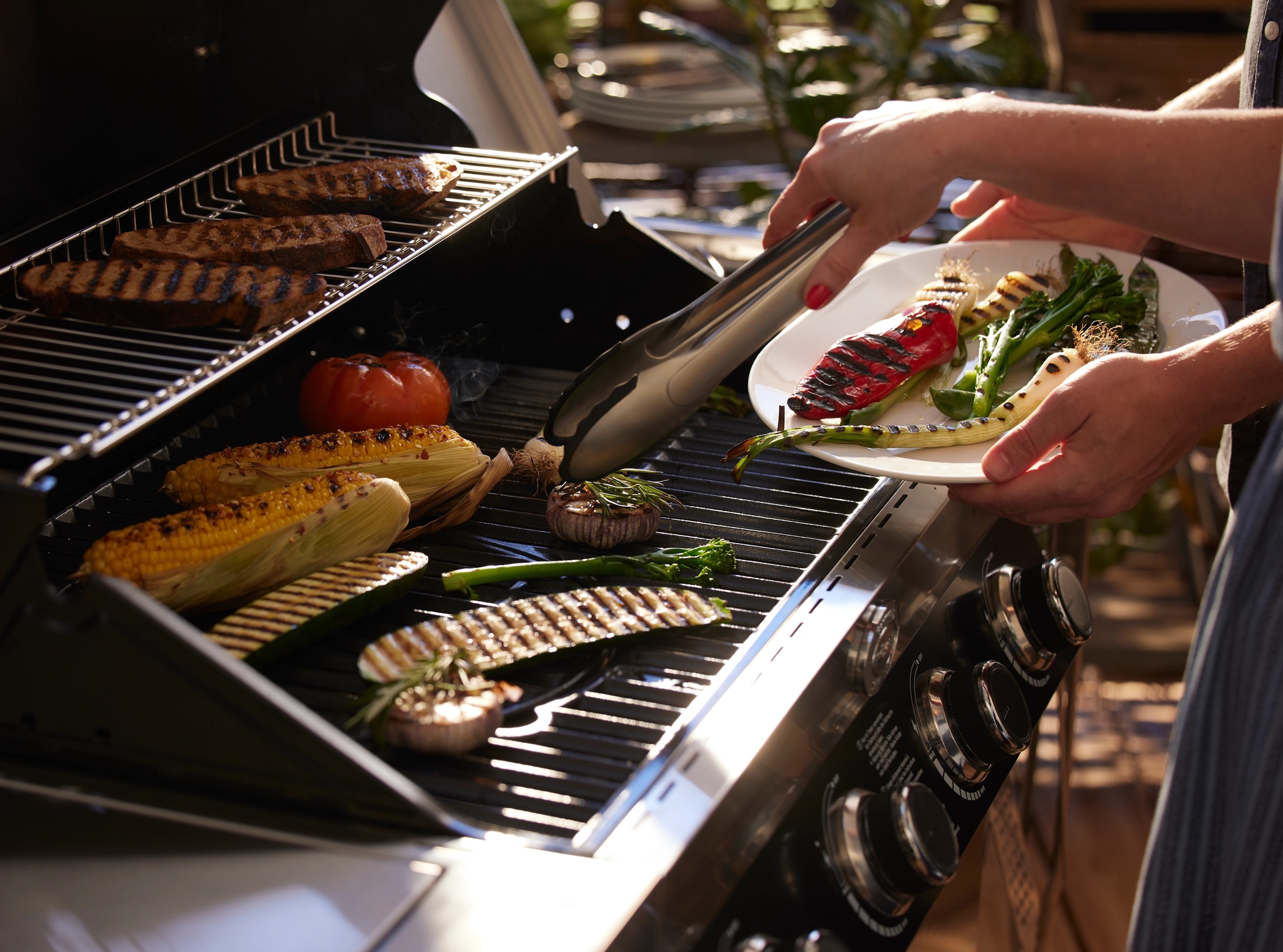 En person legger grillede grønnsaker på en tallerken fra en grill som tilbereder kjøtt, mais og andre grønnsaker