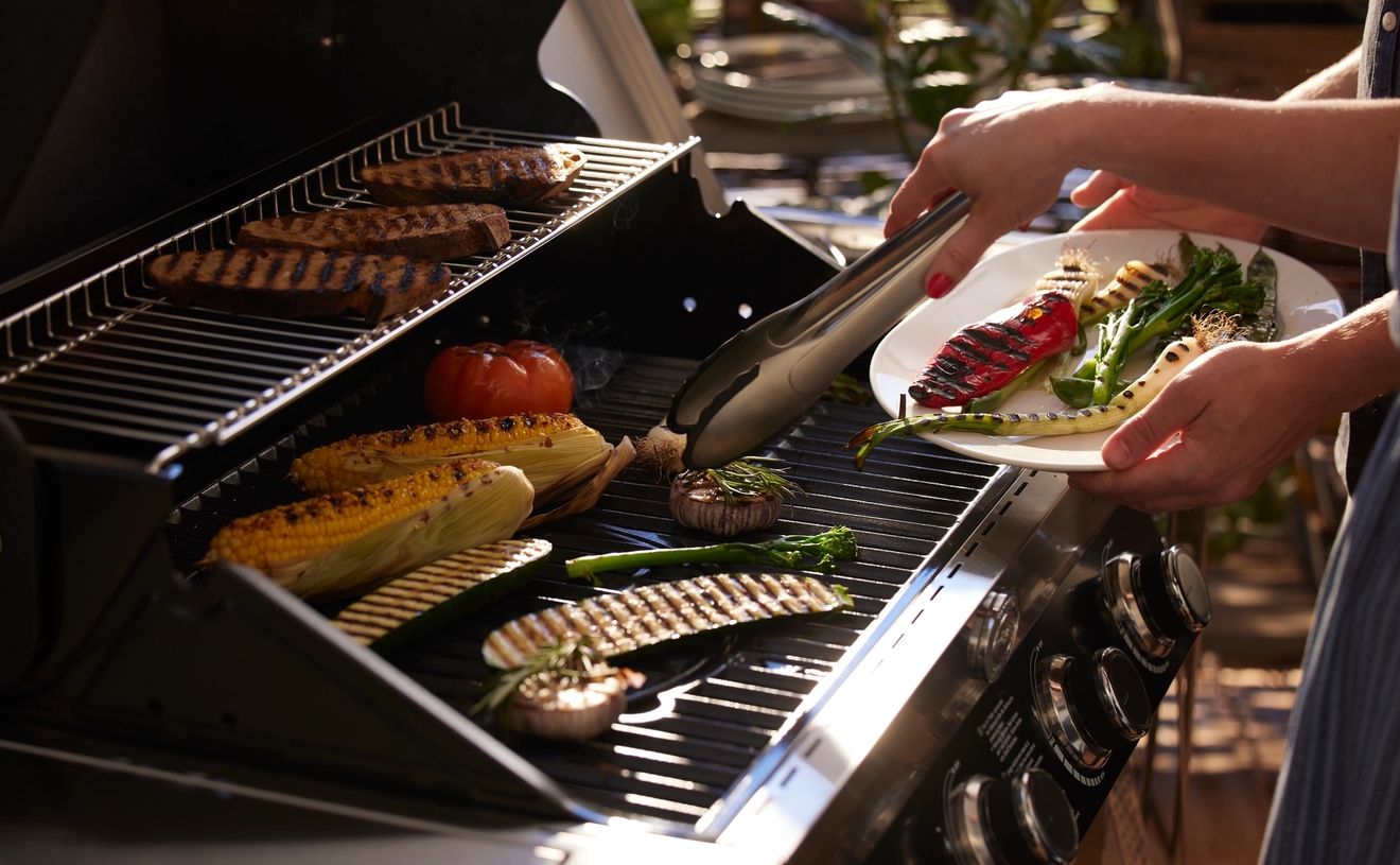 En person lägger grillade grönsaker på en tallrik från en grill där kött, majs och andra grönsaker tillagas