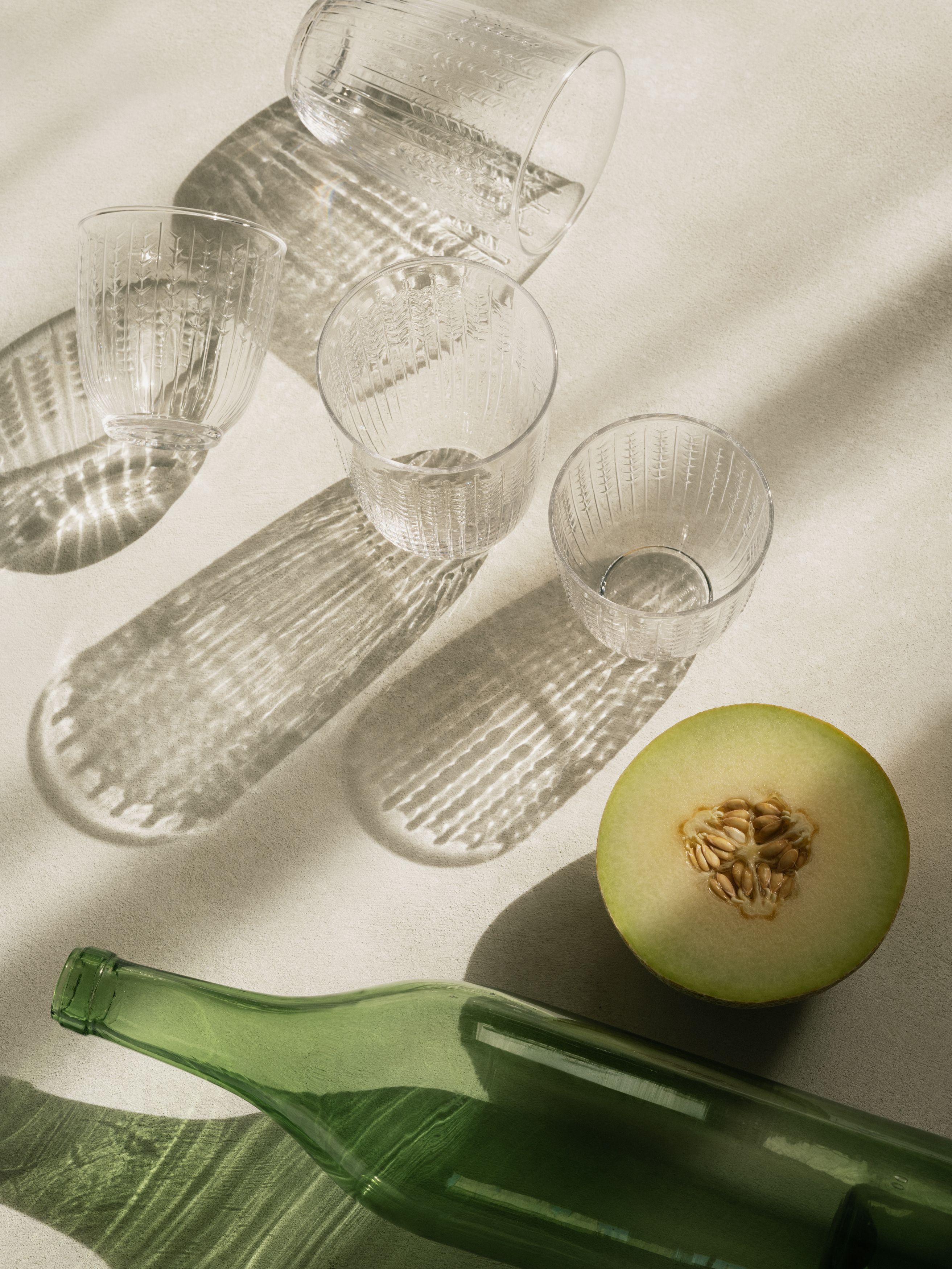 Genomskinliga texturerade glas, en grön flaska och en skivad melon som kastar starka skuggor på en ljus yta.