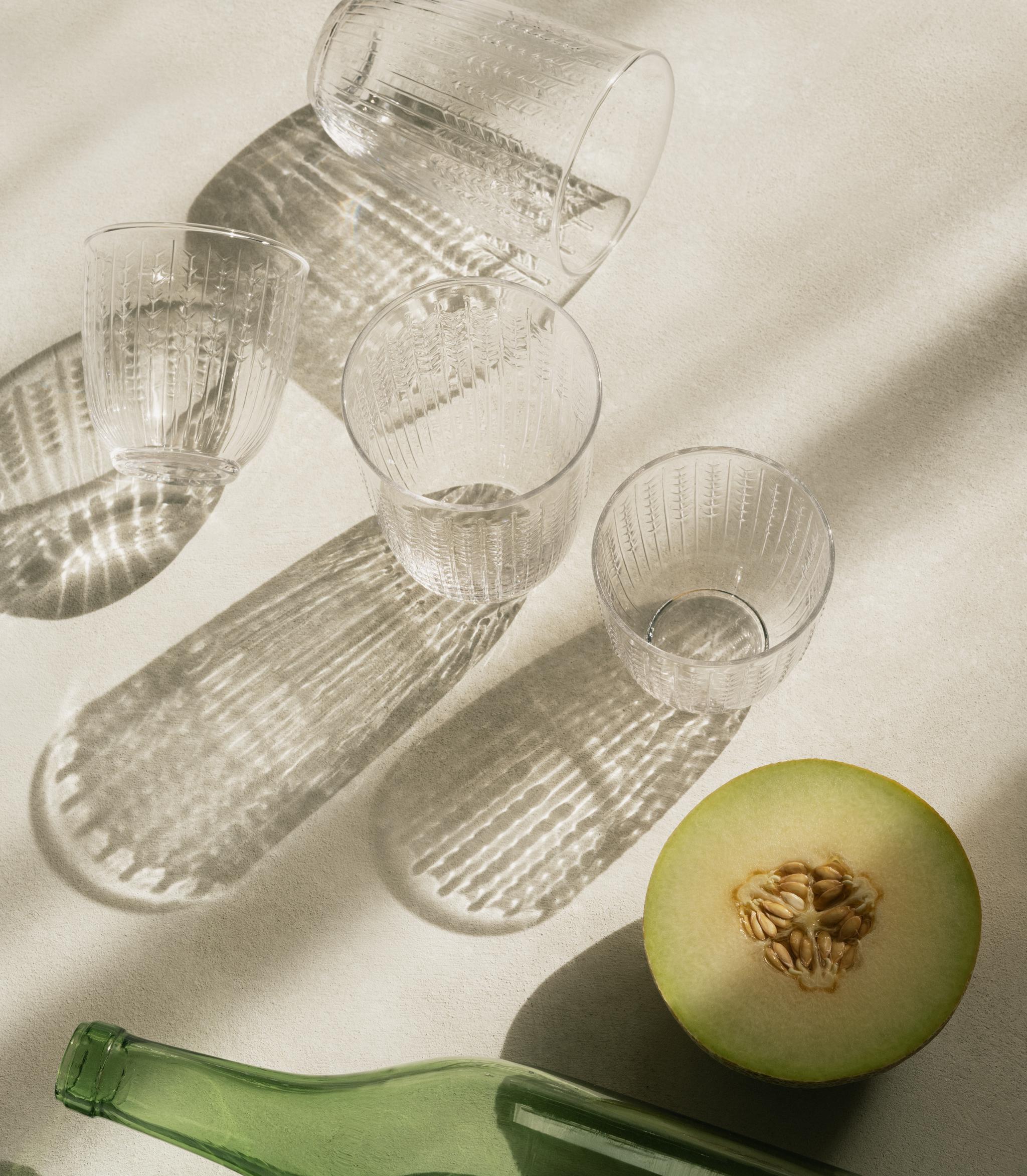 Genomskinliga texturerade glas, en grön flaska och en skivad melon som kastar starka skuggor på en ljus yta.