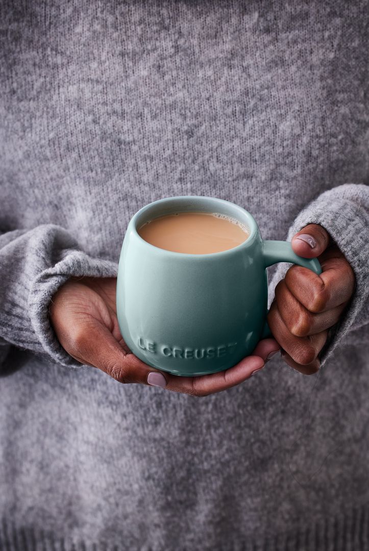 Person i grå stickad tröja håller en turkos Le Creuset-mugg med varm kaffe eller te.