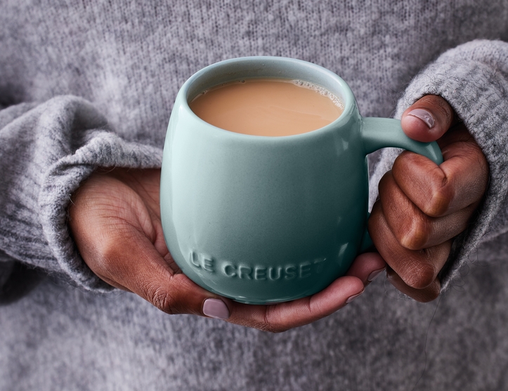 Person i grå stickad tröja håller en turkos Le Creuset-mugg med varm kaffe eller te.