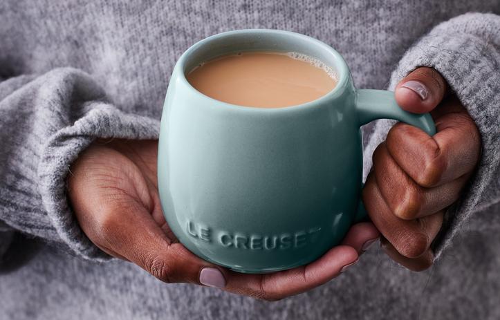 Person i grå stickad tröja håller en turkos Le Creuset-mugg med varm kaffe eller te.