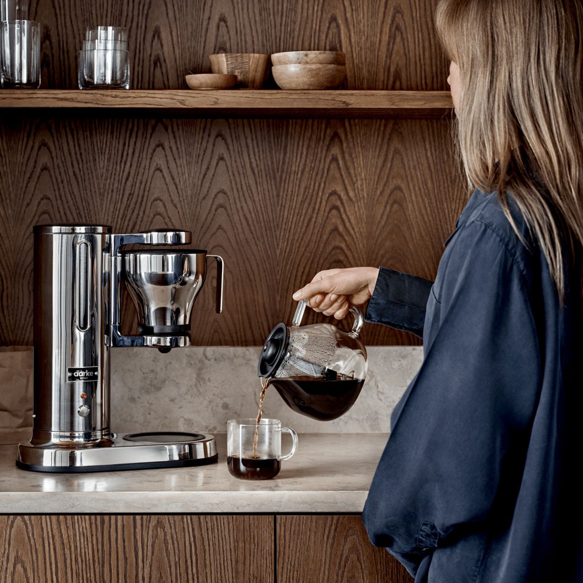 Kaffebryggare i rostfritt stål från Aarke står på en köksbänk och en kvinna häller upp en kopp kaffe