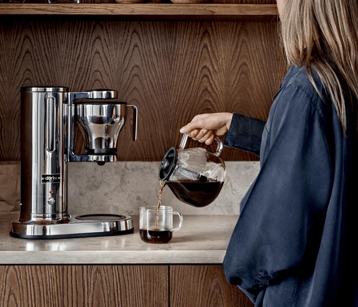 Kaffebryggare i rostfritt stål från Aarke står på en köksbänk och en kvinna häller upp en kopp kaffe