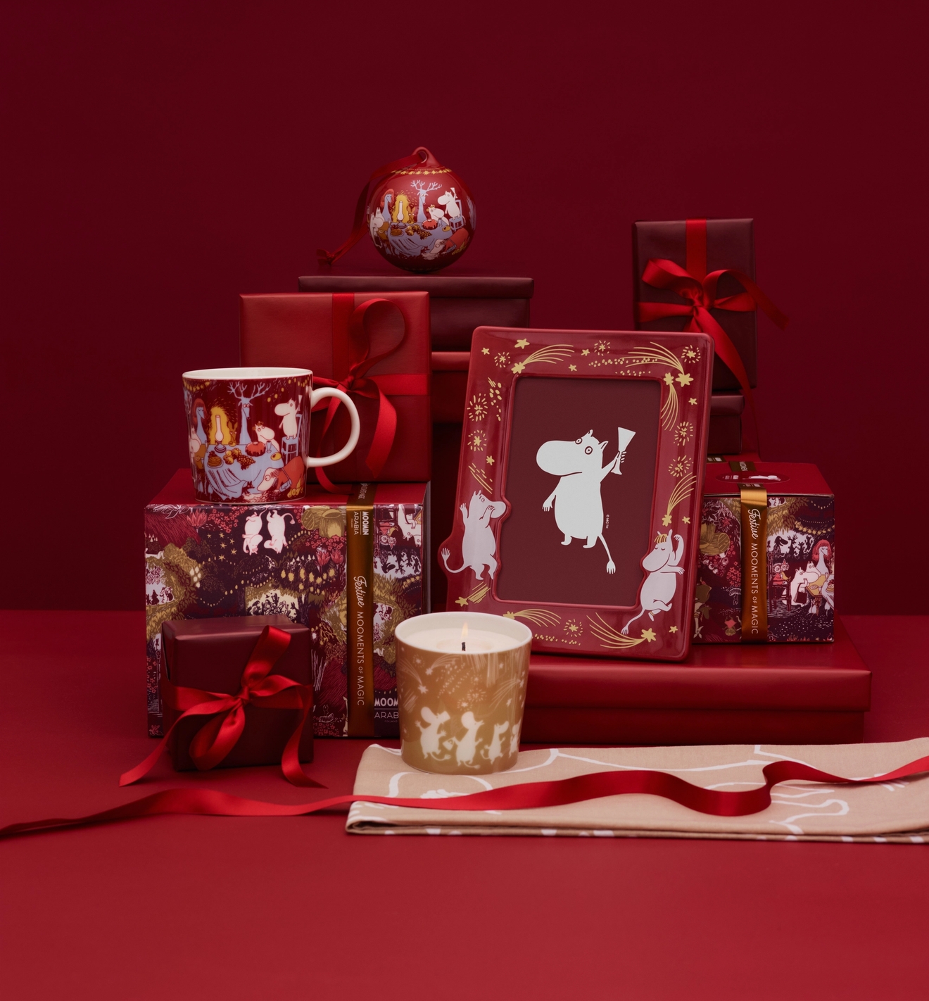 Festive Moomin-themed gifts and decorations in red and gold, including a mug, frame, candle, and gift boxes, on a red background.