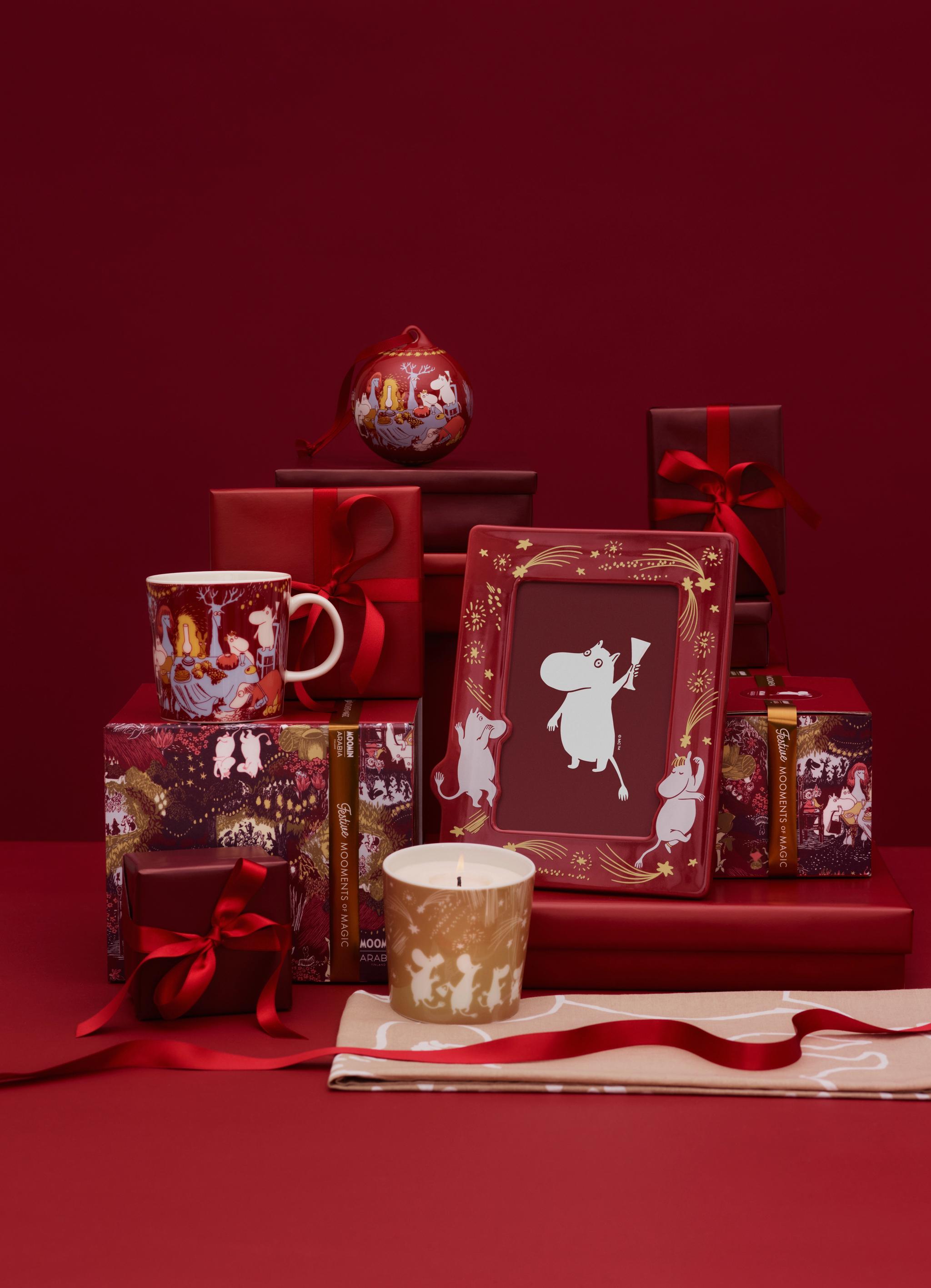 Muumi-aiheisia joululahjoja, kuten muki, kynttilä ja kehystetty kuva, ympäröitynä punaisilla lahjarasioilla ja nauhoilla punaisella taustalla.