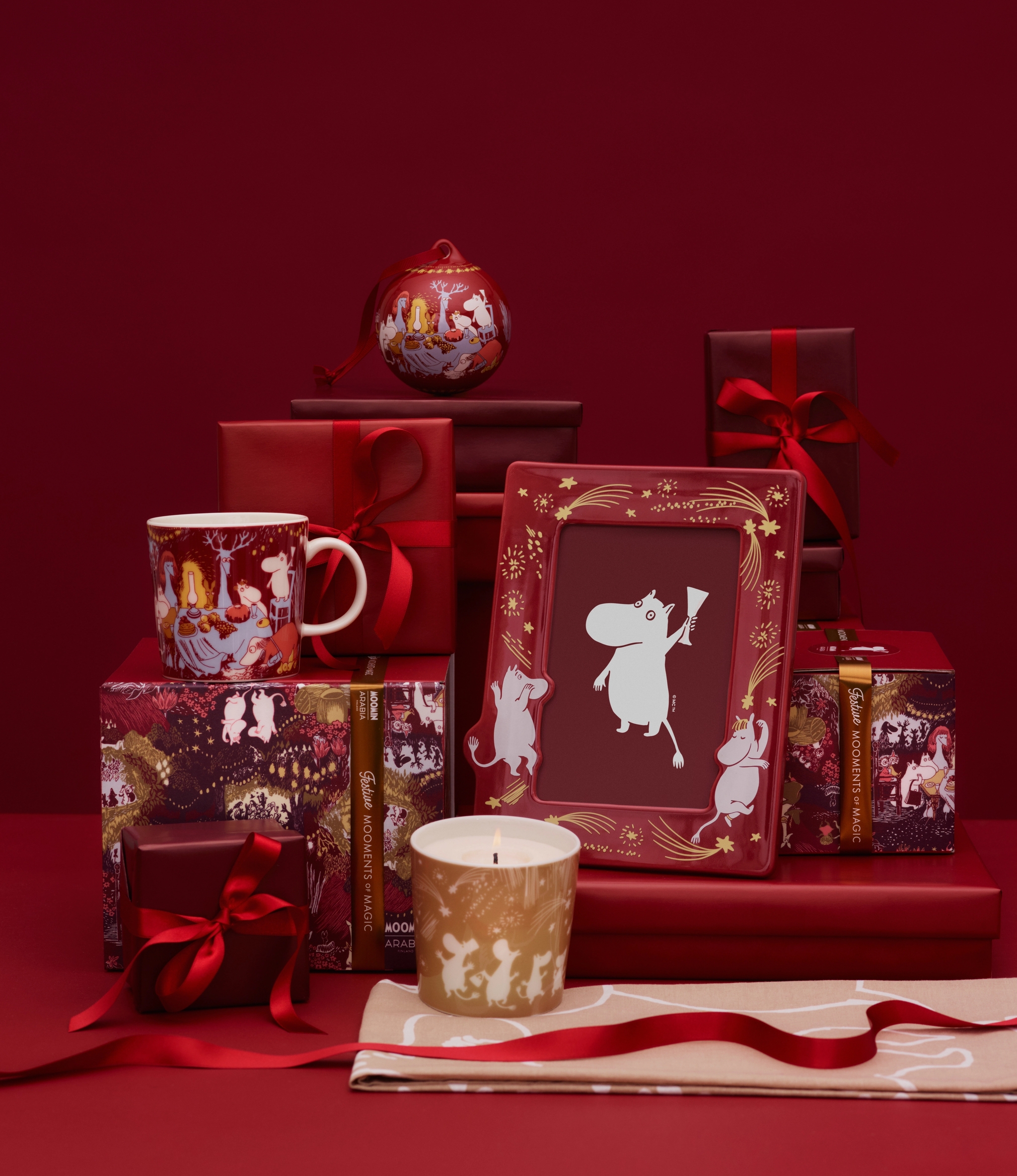 Muumi-aiheisia joululahjoja, kuten muki, kynttilä ja kehystetty kuva, ympäröitynä punaisilla lahjarasioilla ja nauhoilla punaisella taustalla.