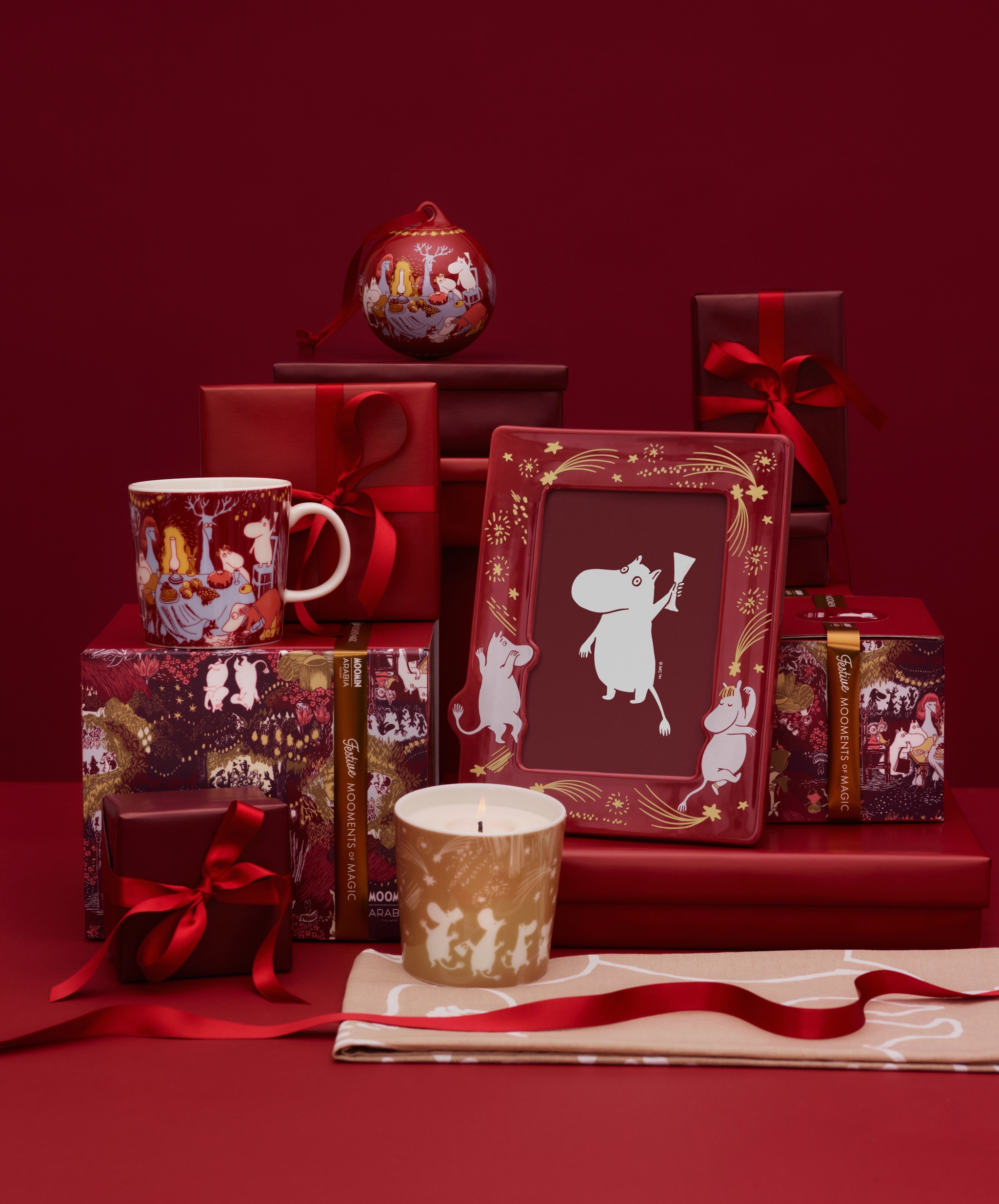 Julklappar med Mumintema, inklusive en mugg, ett ljus och en inramad bild, omgivna av röda presentaskar och band mot en röd bakgrund.