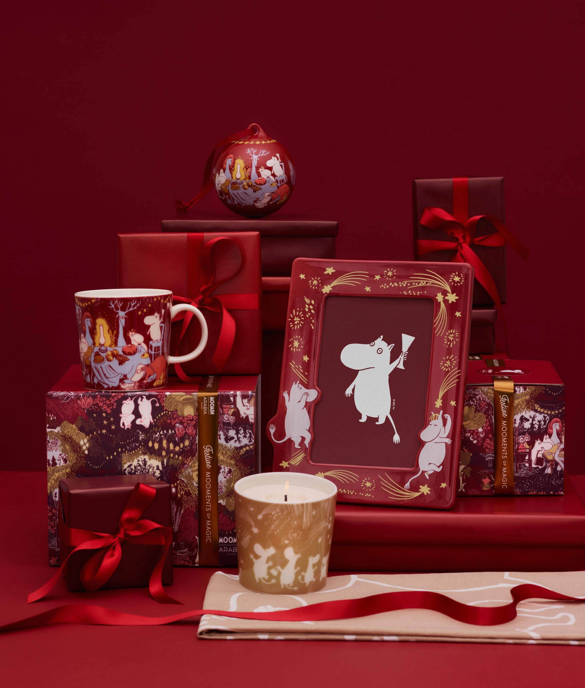 Julklappar med Mumintema, inklusive en mugg, ett ljus och en inramad bild, omgivna av röda presentaskar och band mot en röd bakgrund.