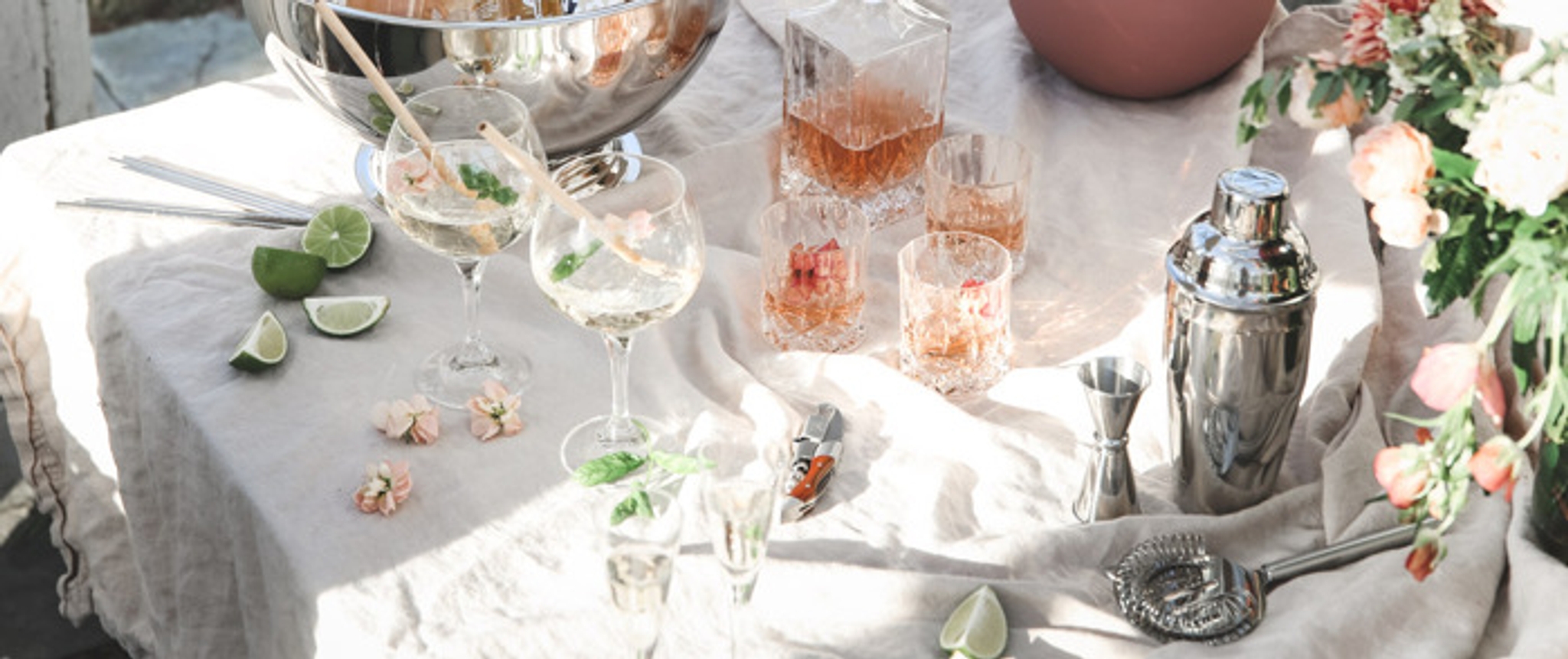Drinktilbehør på bord med hvit linduk og rosa drinker og blomster samt en shaker 