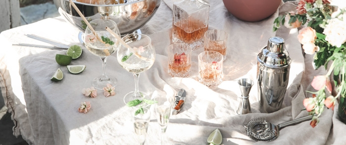 Drinktilbehør på bord med hvit linduk og rosa drinker og blomster samt en shaker