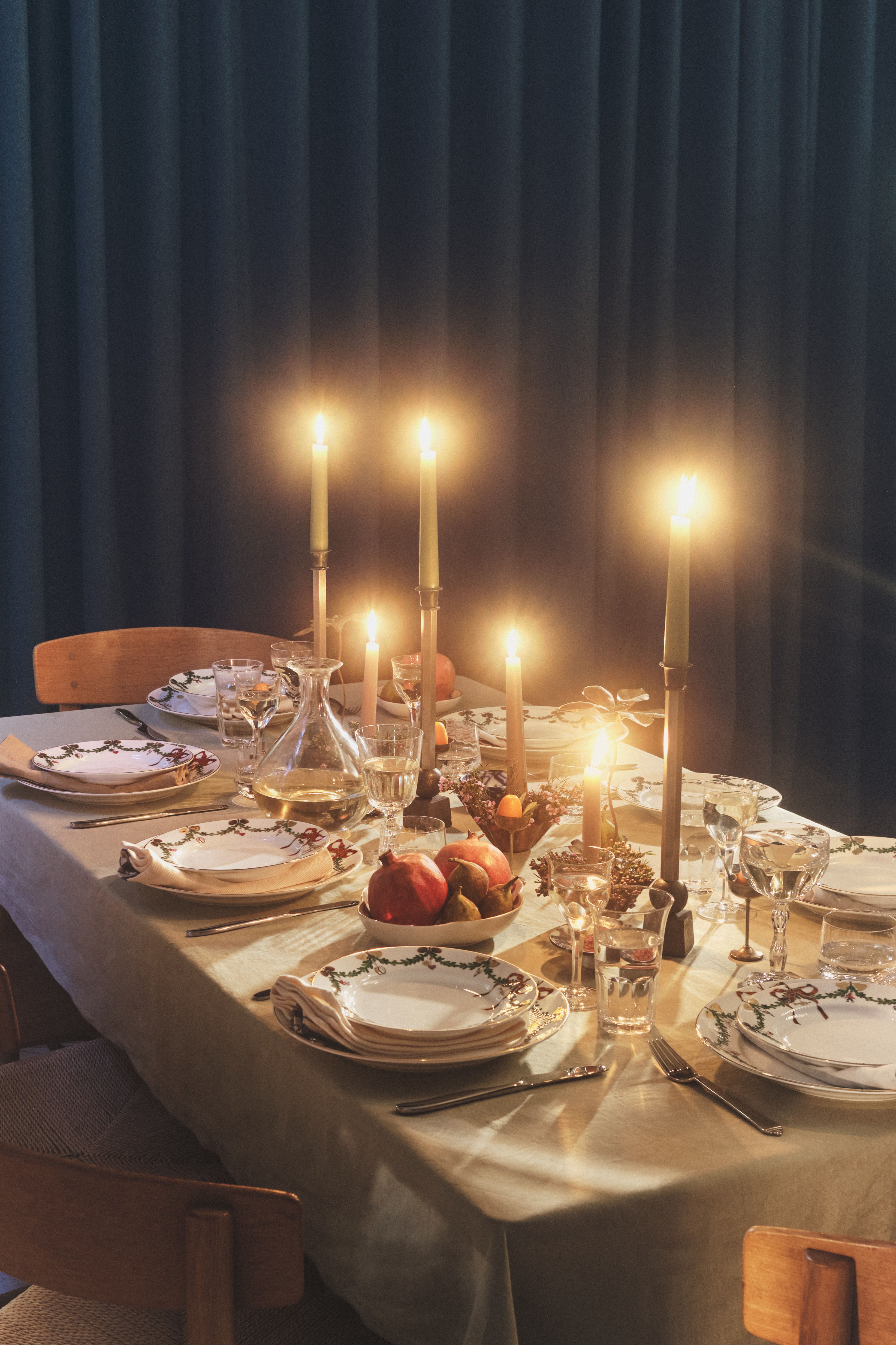 Spisebord med levende lys, dekorative tallerkener, en vinkaraffel og et midtpunkt med frukt.