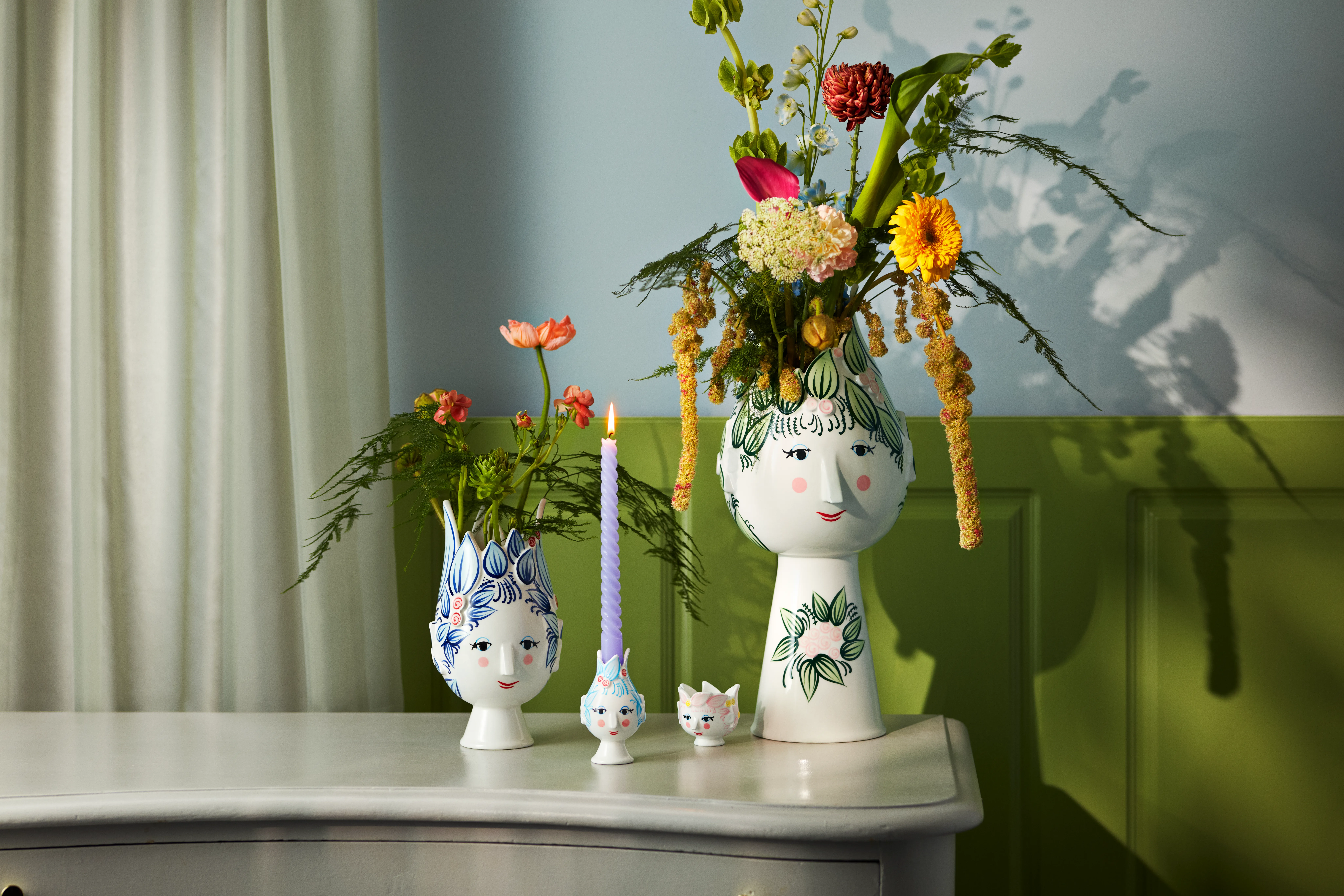 Dekorativa ansiktsformade vaser med blommor, en ljusstake med ett tänt ljus och en liten kattfigur på en byrå