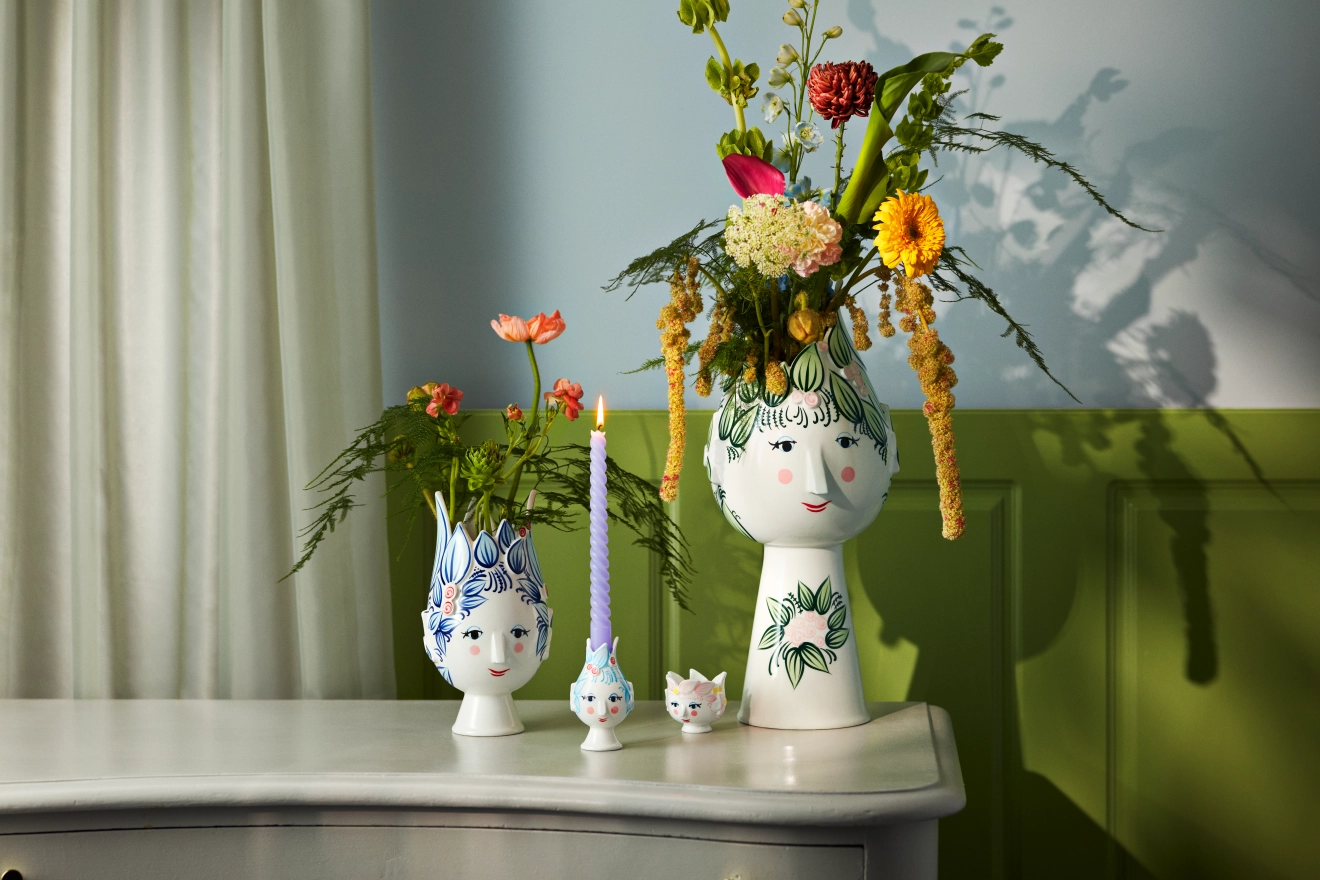 Två stiliserade vaser med blommor, en matchande ljushållare med ett tänt lila ljus och en liten kattfigur står på en byrå mot en ljusblå och grön vägg.