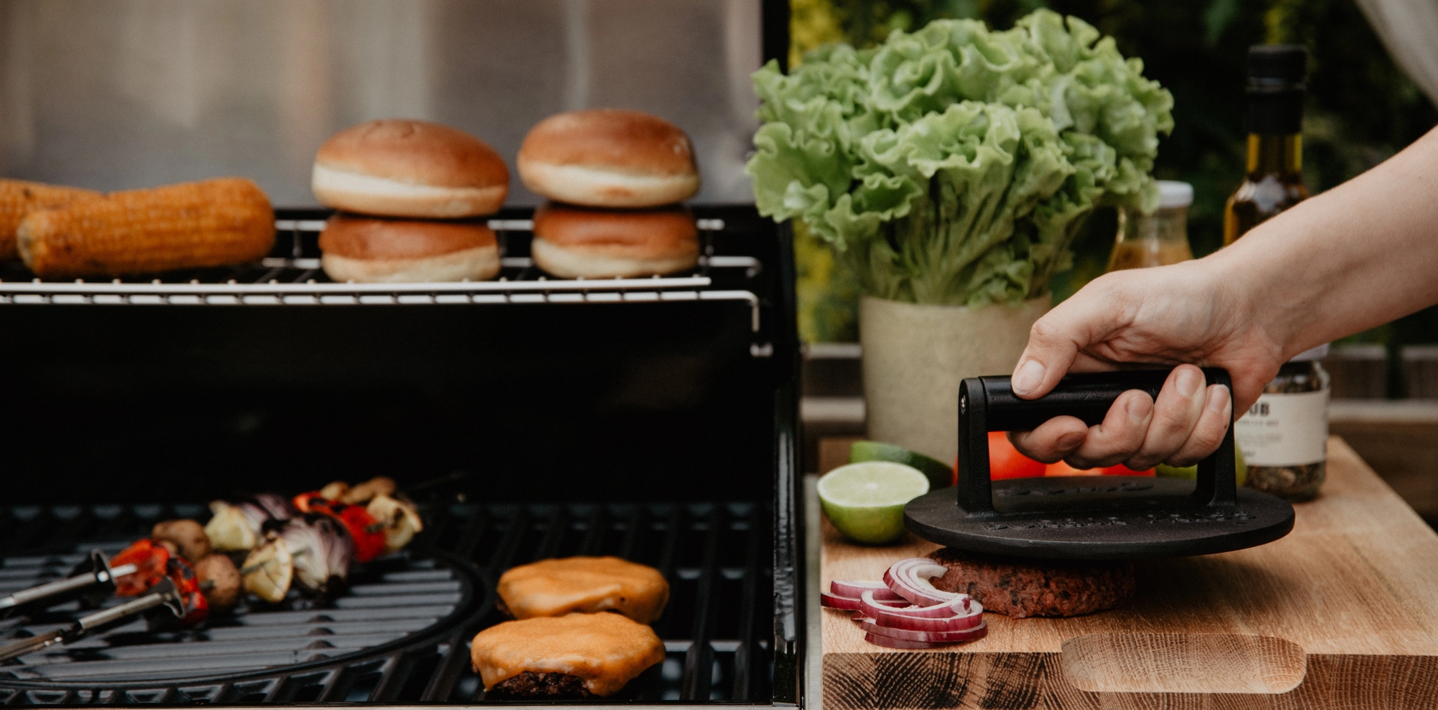 Henkilö prässää hampurilaispihviä puulaudalla kaasugrillin vieressä maissin, sämpylöiden ja kasvisvartaiden kanssa.