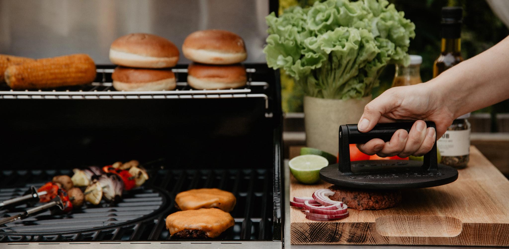 Henkilö prässää hampurilaispihviä puulaudalla kaasugrillin vieressä maissin, sämpylöiden ja kasvisvartaiden kanssa.