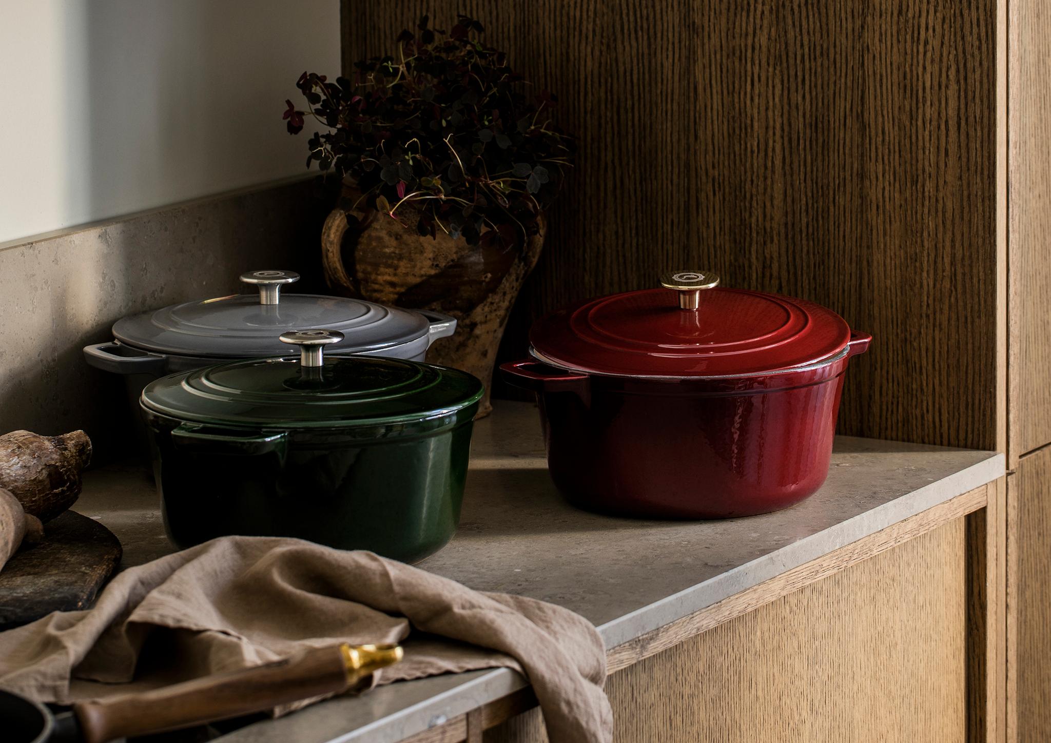 Kolme valurautaista pataa (punainen, vihreä, harmaa) keittiön työtasolla valkoisten ja puisten kaappien alla.