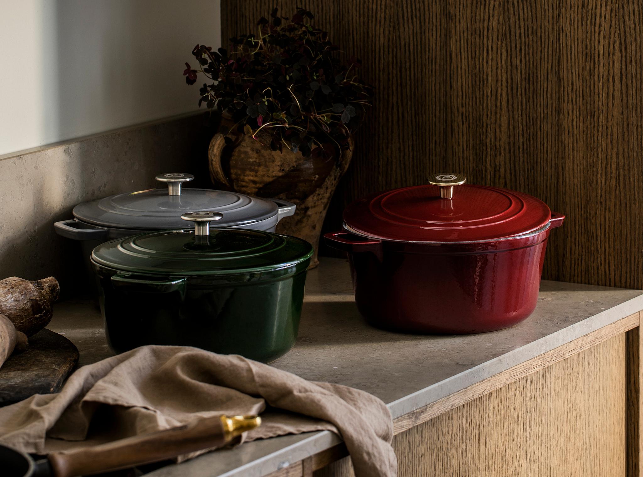 Kolme valurautaista pataa (punainen, vihreä, harmaa) keittiön työtasolla valkoisten ja puisten kaappien alla.