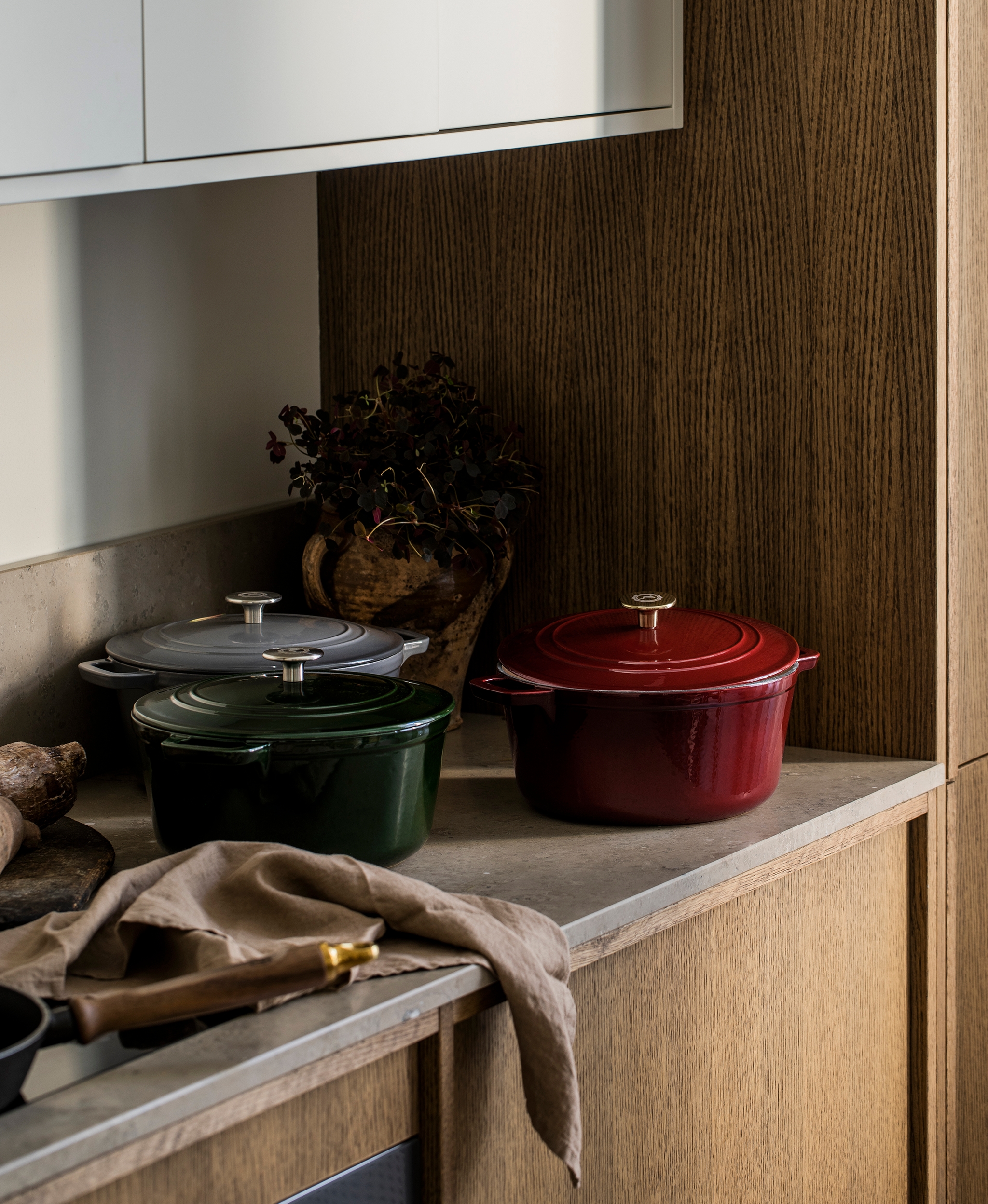 Kolme valurautaista pataa (punainen, vihreä, harmaa) keittiön työtasolla valkoisten ja puisten kaappien alla.