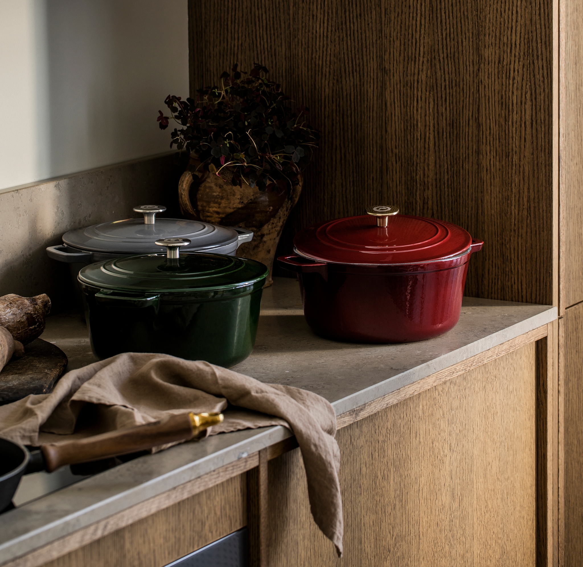 Kolme valurautaista pataa (punainen, vihreä, harmaa) keittiön työtasolla valkoisten ja puisten kaappien alla.