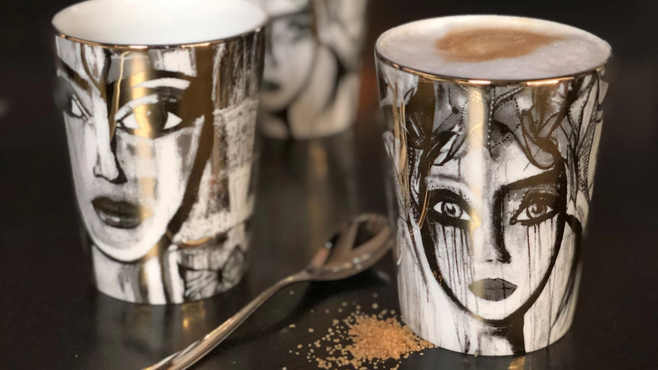 Taidekuvioidut kahvimukit, joissa on mustavalkoisia kasvojen piirroksia, pöydällä lusikan ja sokerirakeiden kanssa.