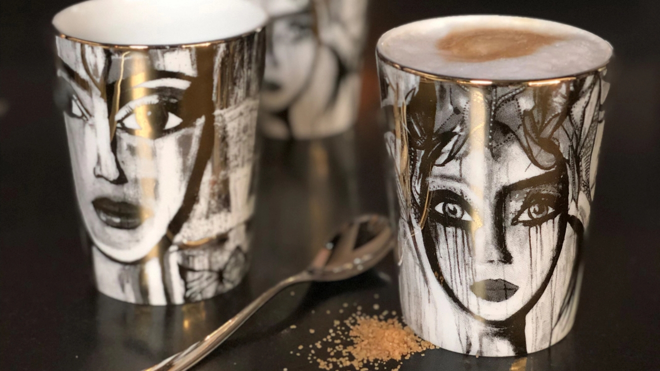 Taidekuvioidut kahvimukit, joissa on mustavalkoisia kasvojen piirroksia, pöydällä lusikan ja sokerirakeiden kanssa.