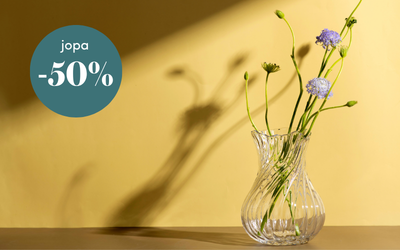 Keltainen mainoskuva, jossa lasimaljakossa on kukkia ja vihreässä ympyrässä teksti ”jopa -50 %” alennuksesta.