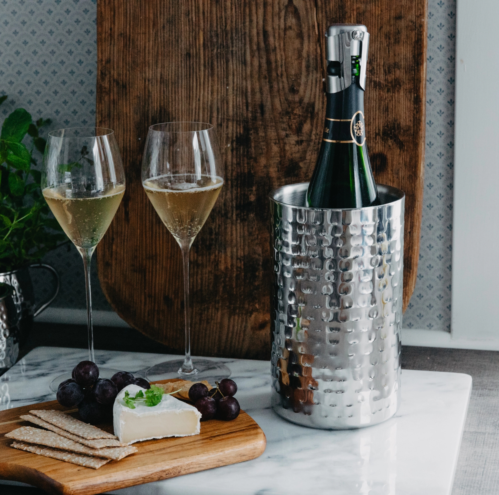 En champagneflaska i en silverfärgad vinkylare, två glas mousserande vin och en ostbricka med brie, vindruvor och kex.