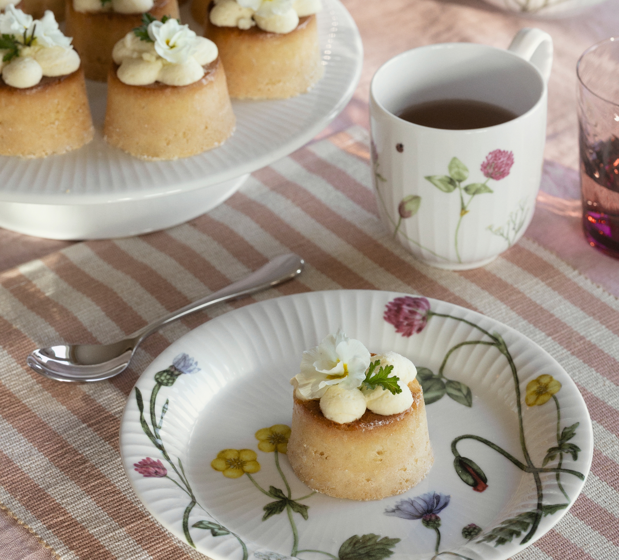 Dukad fikabord med små bakelser med grädde och ätbara blommor, te, jordgubbar och citroner på blommigt porslin.