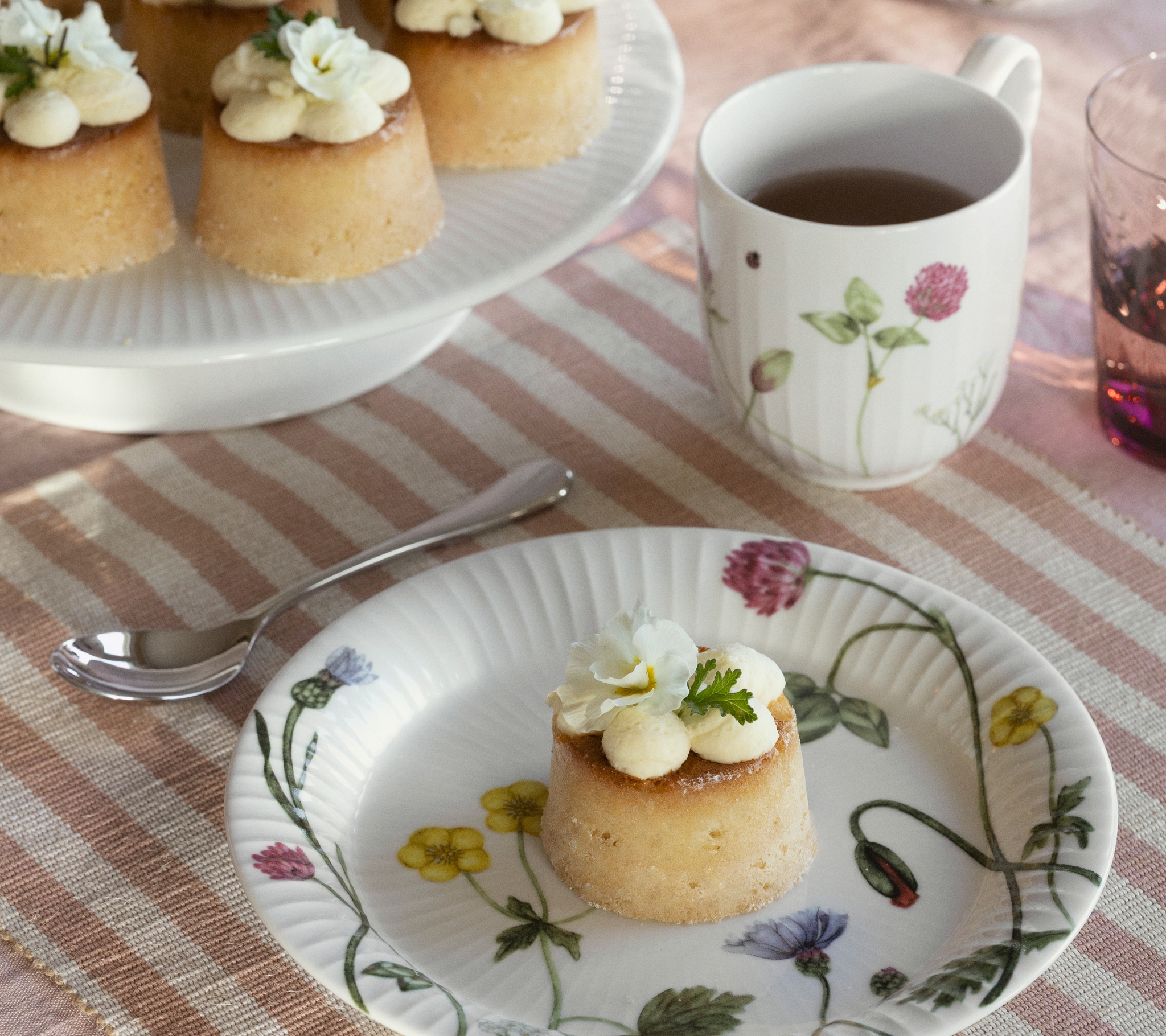 Ett utomhusbord dukat med små kakor toppade med vita blommor, färska jordgubbar, citroner och en blommig mugg te på en randig duk.
