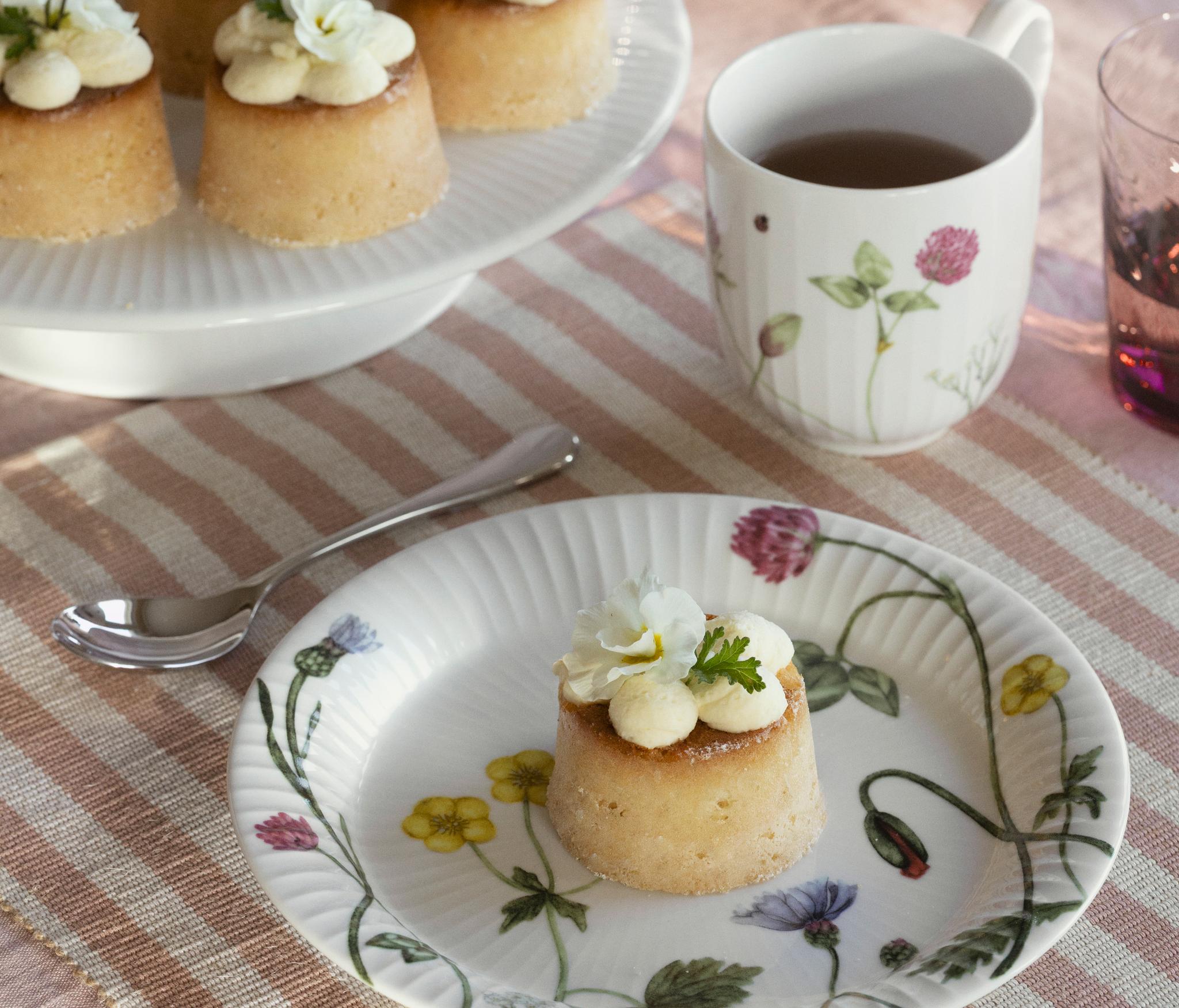 Ett utomhusbord dukat med små kakor toppade med vita blommor, färska jordgubbar, citroner och en blommig mugg te på en randig duk.