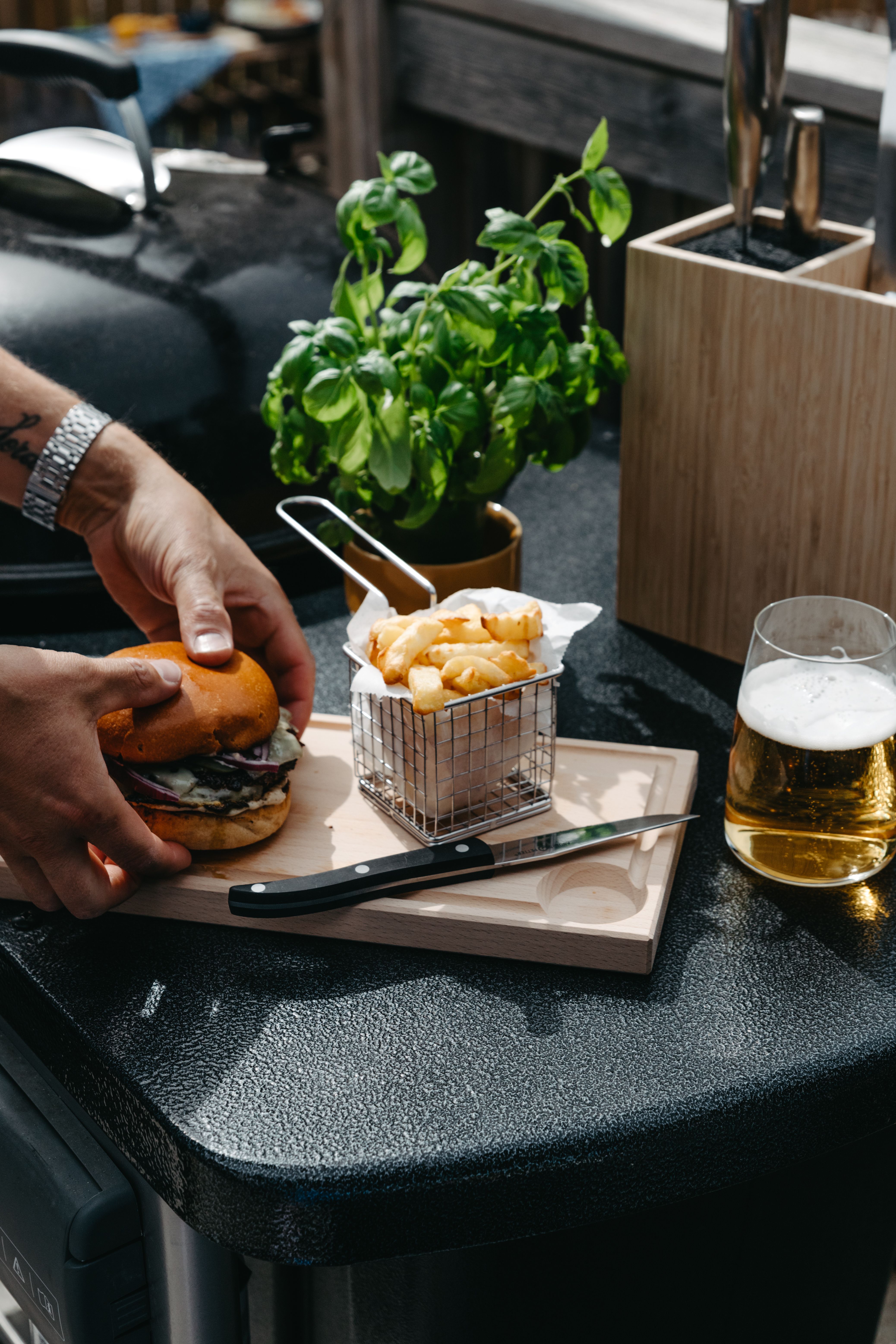Person som förbereder en grillad hamburgare med pommes frites och öl på en uteplats.