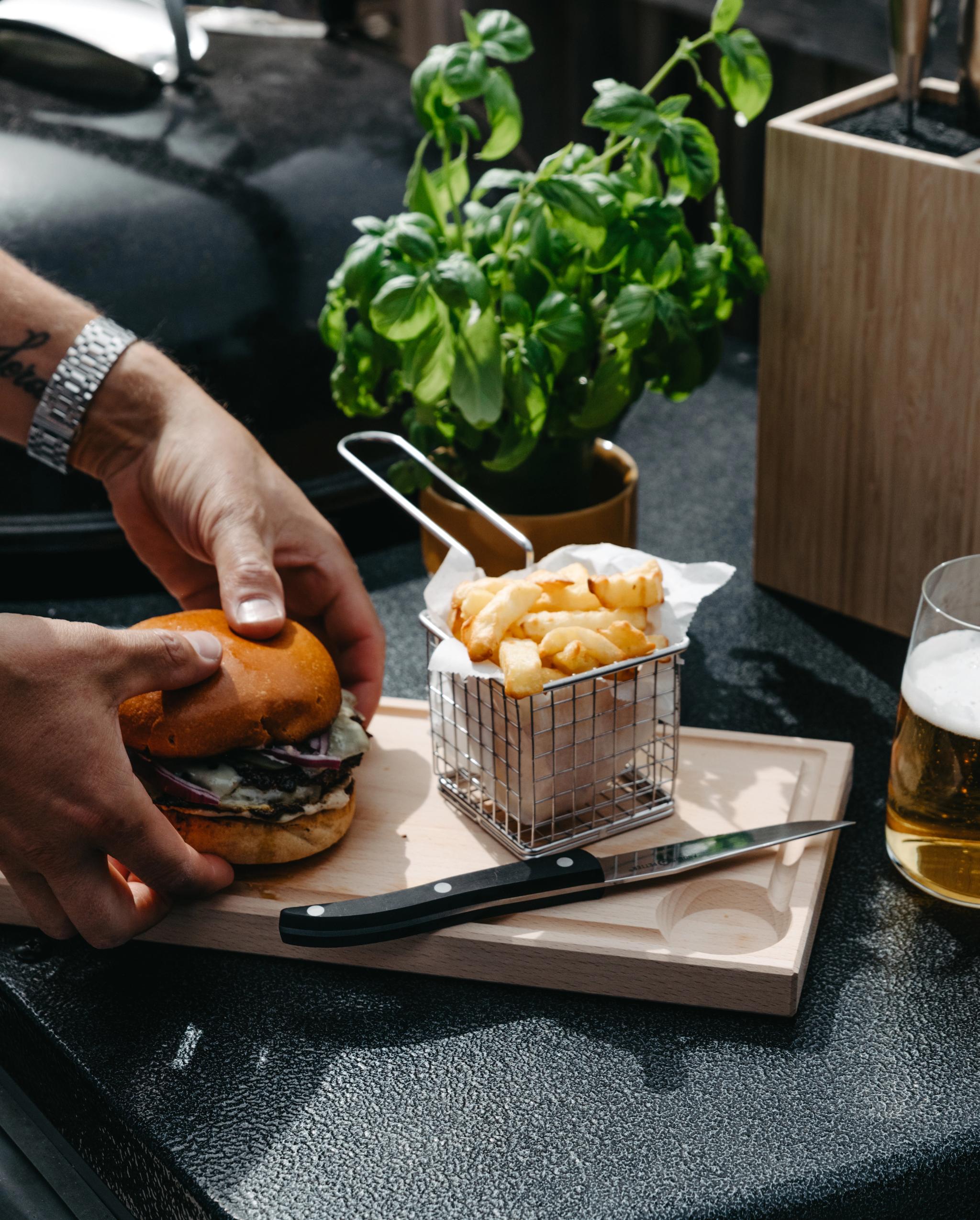 Person som förbereder en grillad hamburgare med pommes frites och öl på en uteplats.