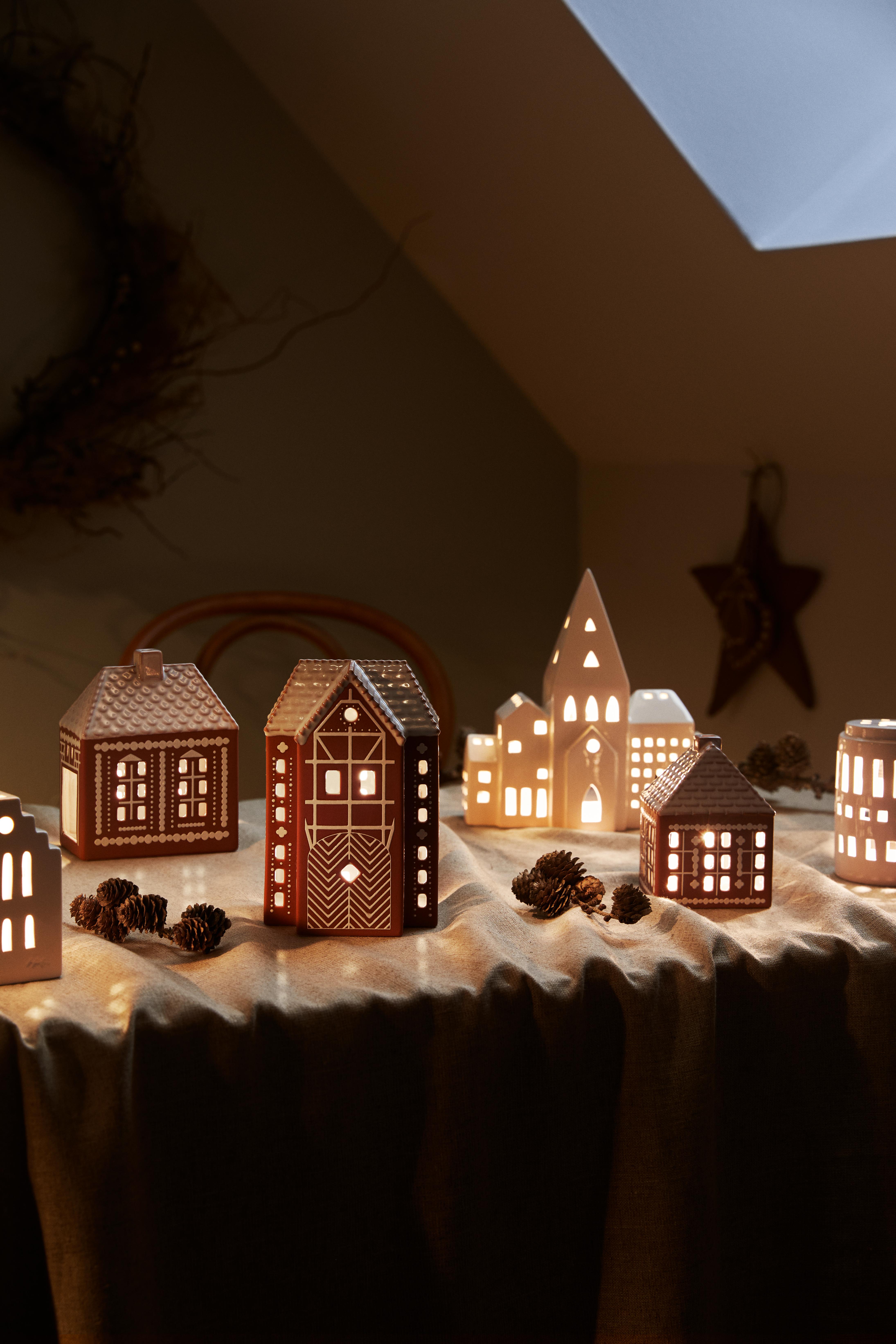A cozy display of glowing ceramic house luminaries in white and gingerbread-like designs, arranged on a table with pinecones.