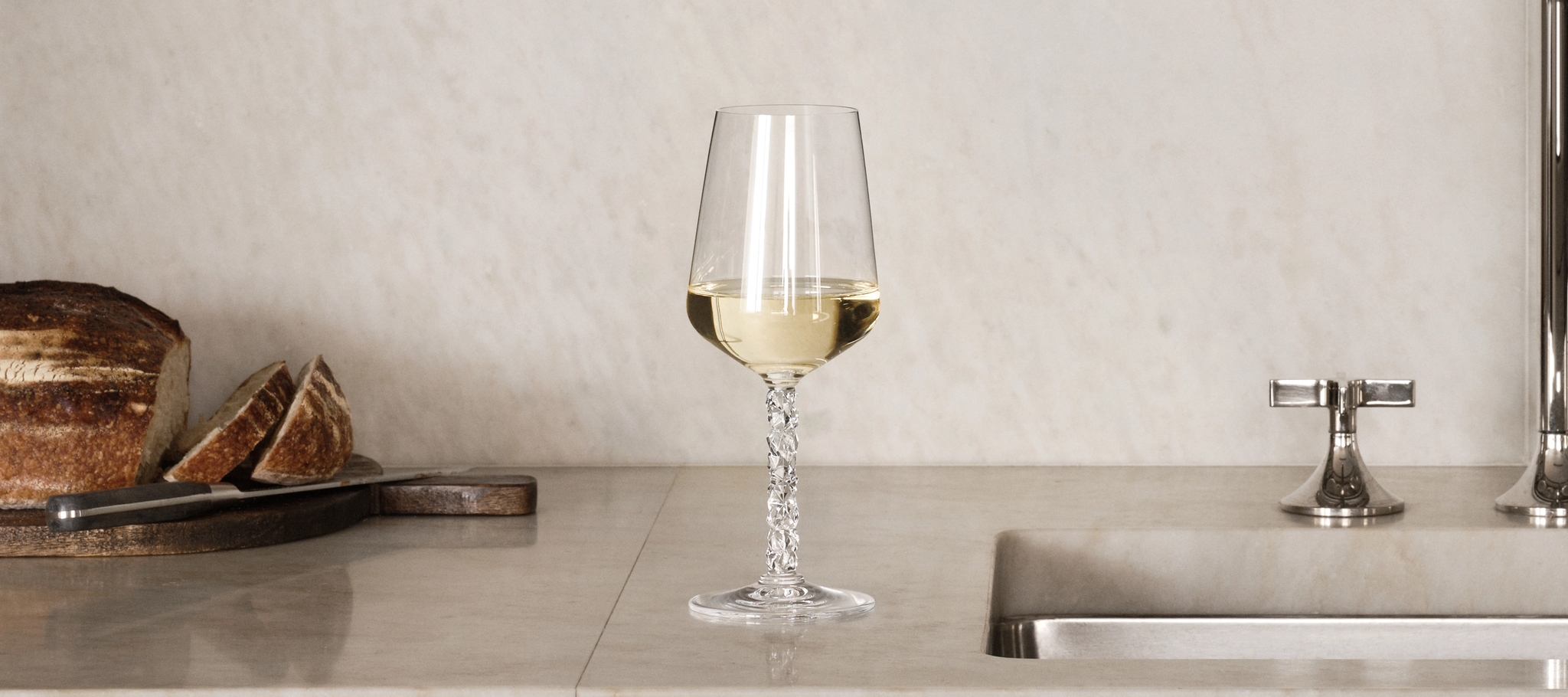 Elegant marmorkök med öppna hyllor, kristallglas, vitt porslin, bröd och ett glas vitt vin vid diskhon.