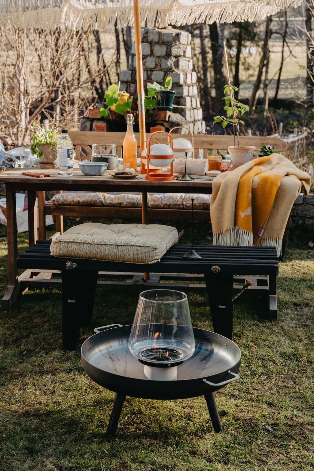 En utomhusscen med ett träbord dukat med mat och dryck, bänkar med kuddar, en gul filt och en modern eldstad med en glasinkapslad låga i förgrunden.