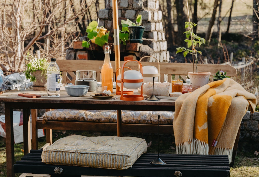 En utomhusscen med ett träbord dukat med mat och dryck, bänkar med kuddar, en gul filt och en modern eldstad med en glasinkapslad låga i förgrunden.