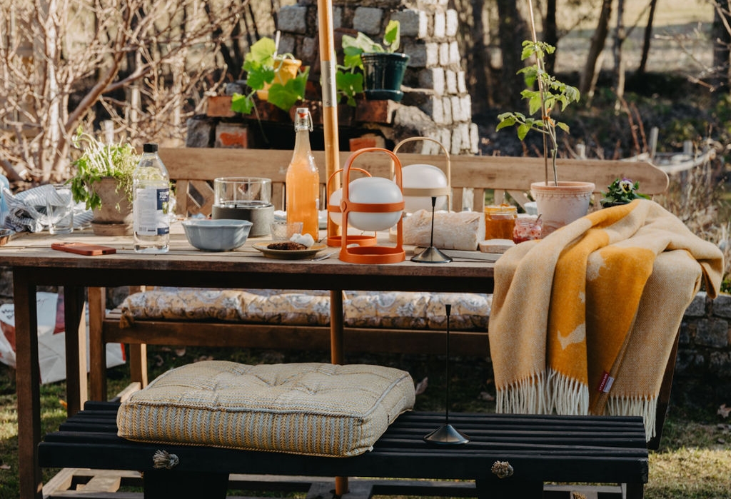 En utomhusscen med ett träbord dukat med mat och dryck, bänkar med kuddar, en gul filt och en modern eldstad med en glasinkapslad låga i förgrunden.
