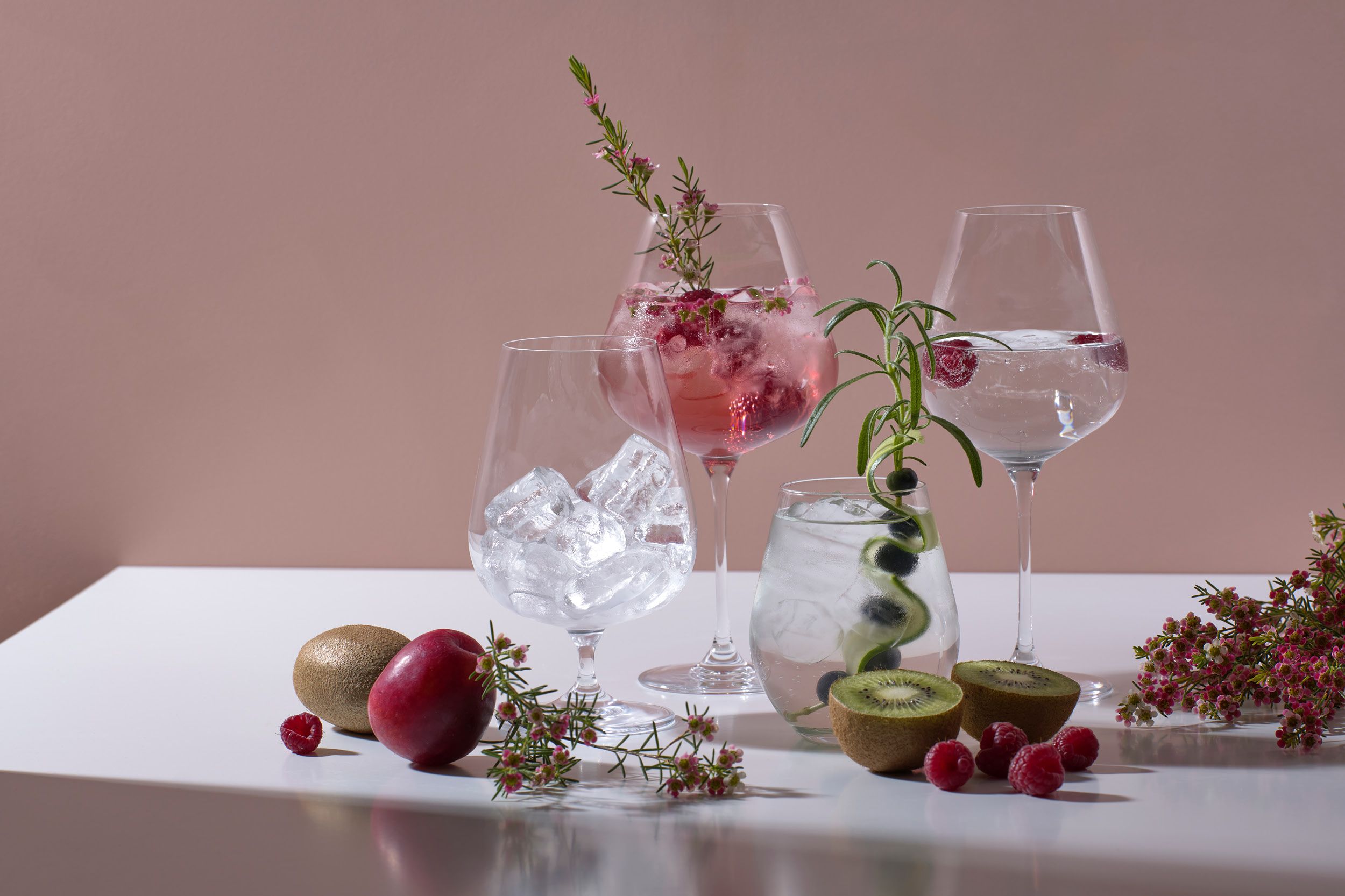 Elegante drinkglass med is, bringebær, kiwi, rosmarin og dekorative blomster på et hvitt bord.