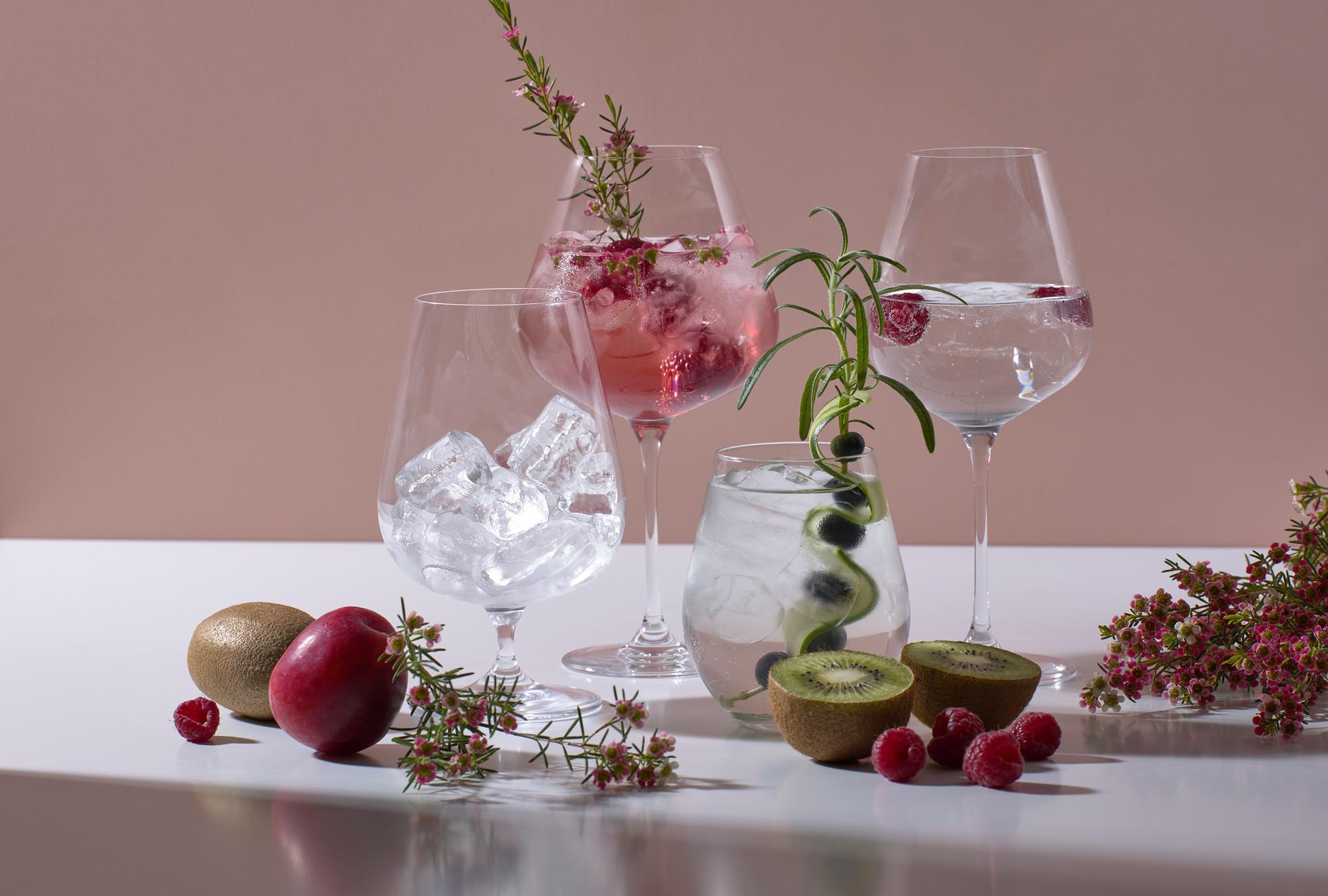 Elegante drinkglass med is, bringebær, kiwi, rosmarin og dekorative blomster på et hvitt bord.