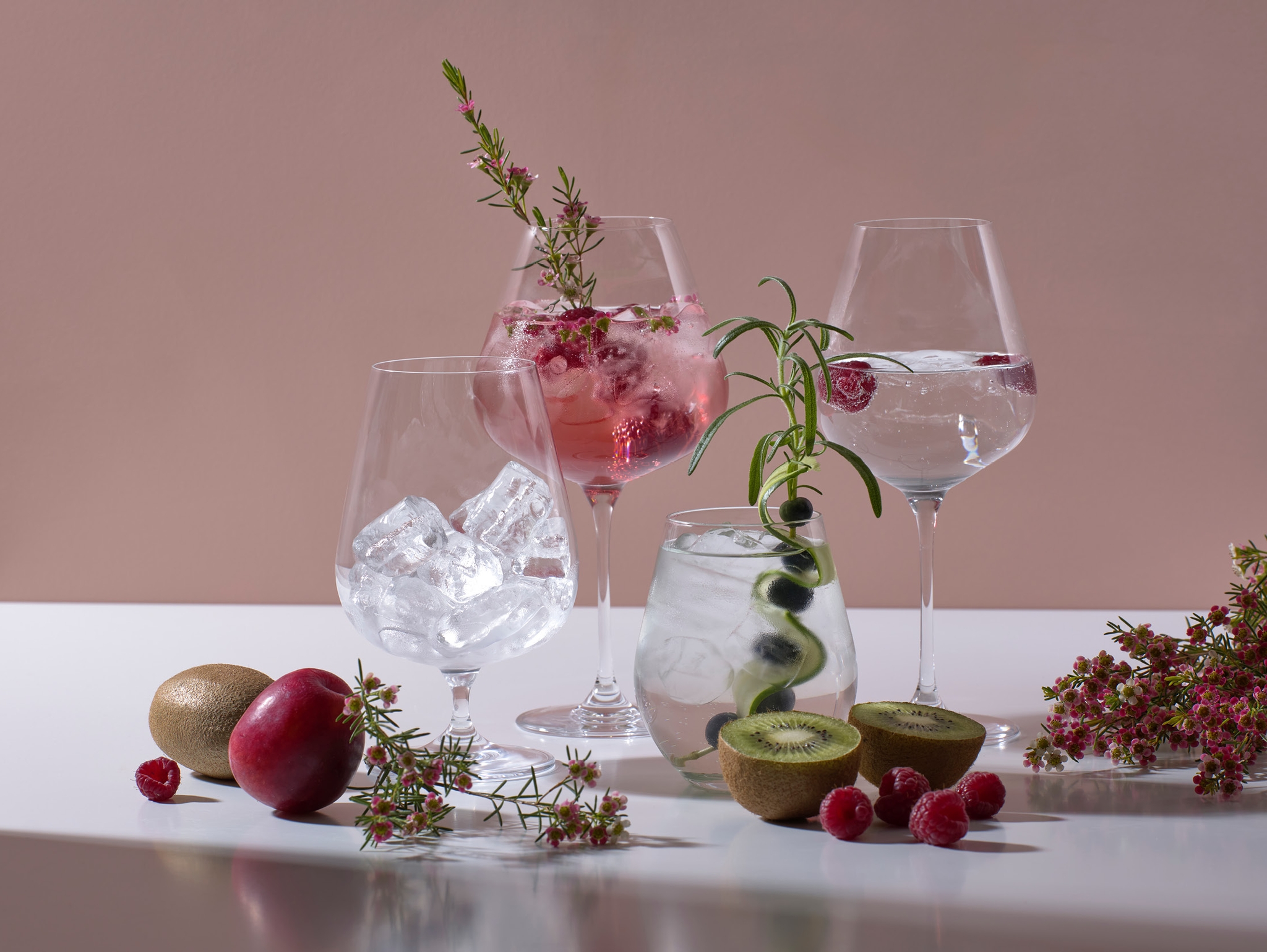 Elegante drinkglass med is, bringebær, kiwi, rosmarin og dekorative blomster på et hvitt bord.