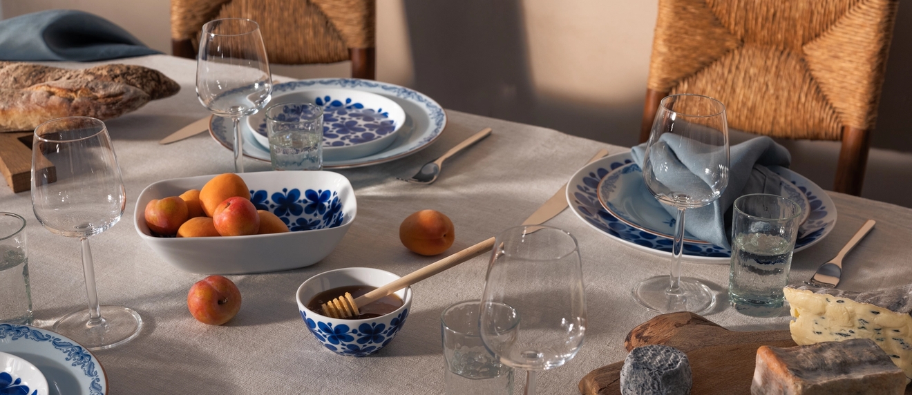 Ett matbord dukat med blåvita tallrikar, vinglas, aprikoser, honung, olika ostar och prosciutto, med halmstolar i bakgrunden.