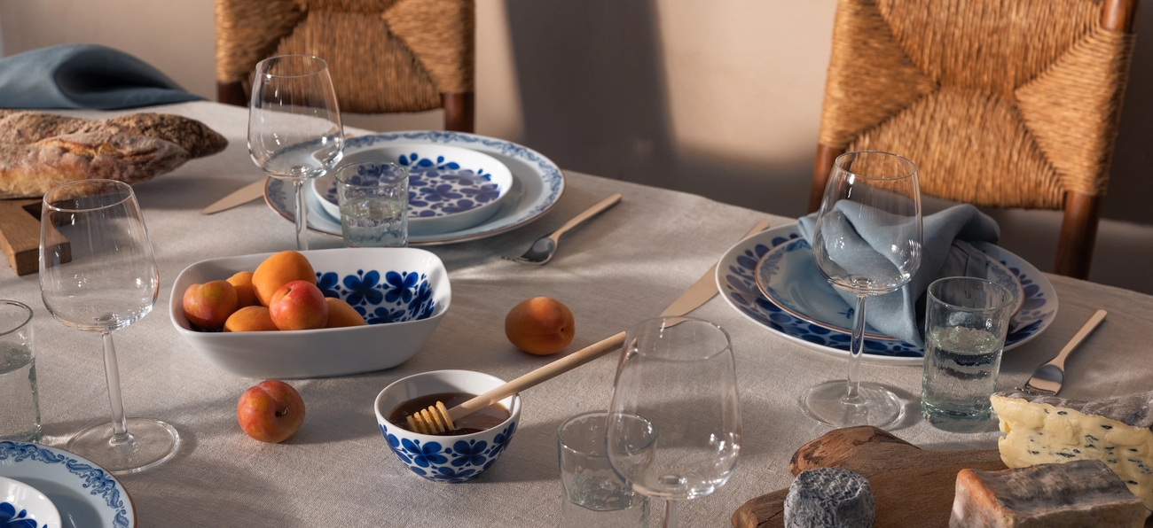 Ett matbord dukat med blåvita tallrikar, vinglas, aprikoser, honung, olika ostar och prosciutto, med halmstolar i bakgrunden.