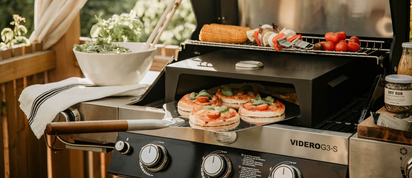 Somrig utomhusgrill med pizzor, grönsaksspett och majs på en modern gasolgrill på altanen.