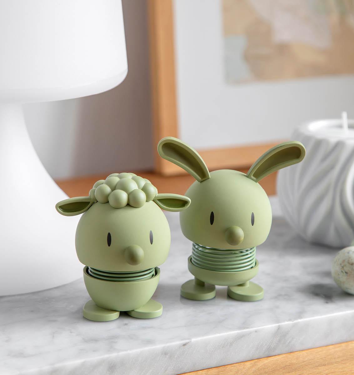 Two light green minimalist spring figures of a sheep and a rabbit stand on a marble surface next to speckled eggs.