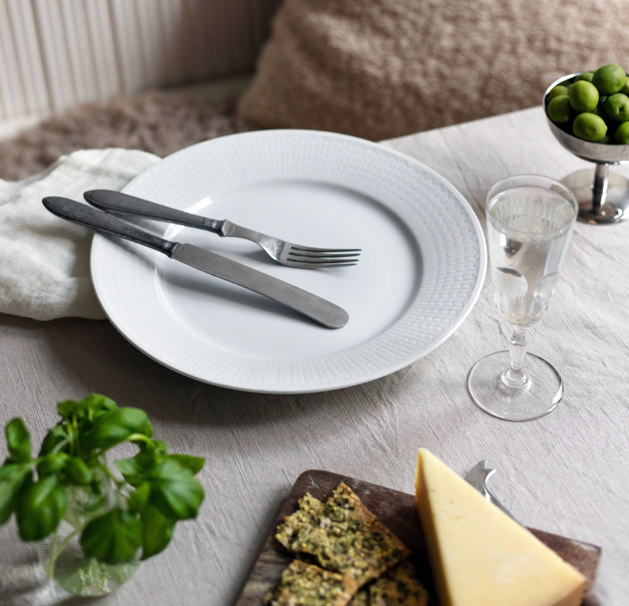 Dekket bord med hvit tallerken, bestikk, grønn oliven, urter og ost i et lyst og hyggelig spisehjørne.
