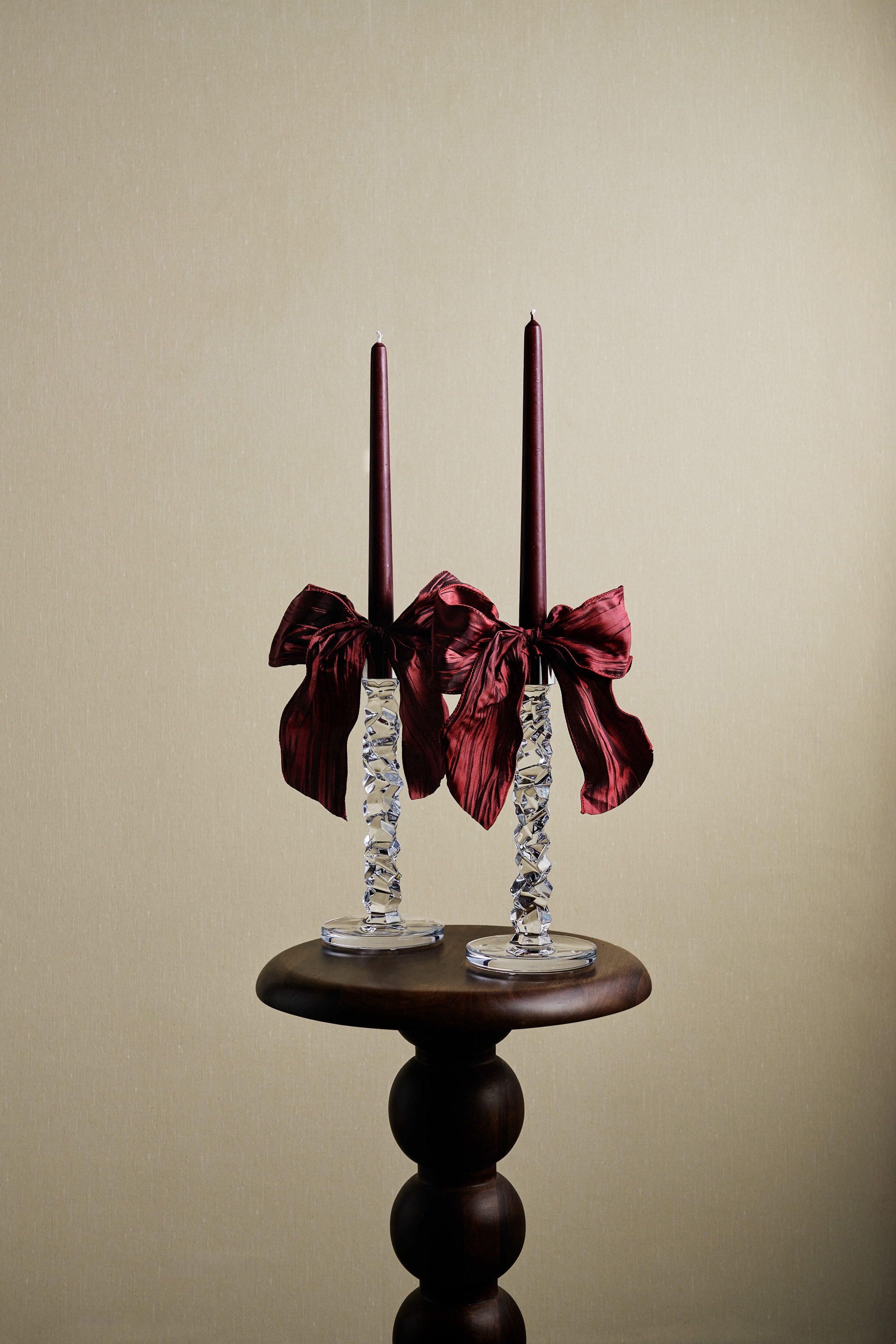 Två mörkröda ljus i fasetterade kristallhållare med matchande rosetter, på ett mörkt träpiedestalbord.