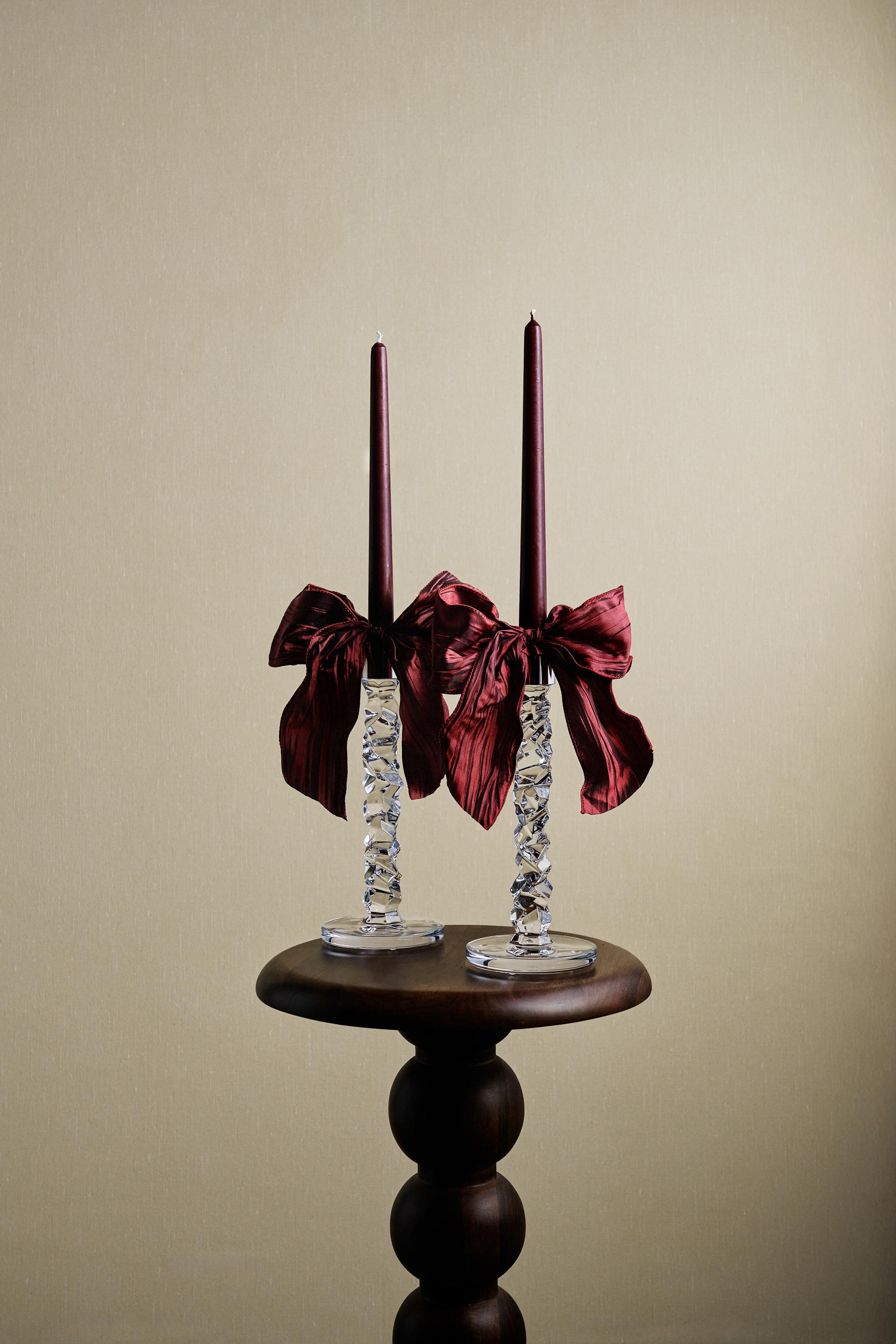 Kaksi tummanpunaista kartiokynttilää teksturoiduissa lasisissa kynttilänjaloissa, jotka on sidottu tummanpunaisella rusetilla, pyöreällä puisella pöydällä.