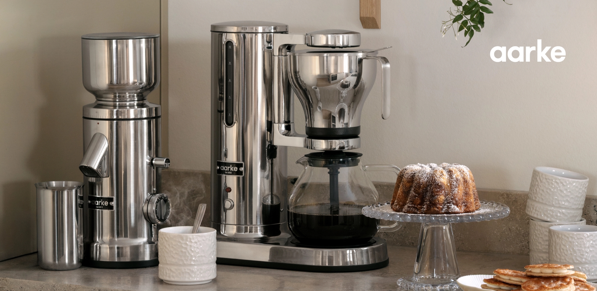 Aarke kaffekvarn och kaffebryggare i rostfritt stål på en köksbänk med en bundtkaka och pannkakor.
